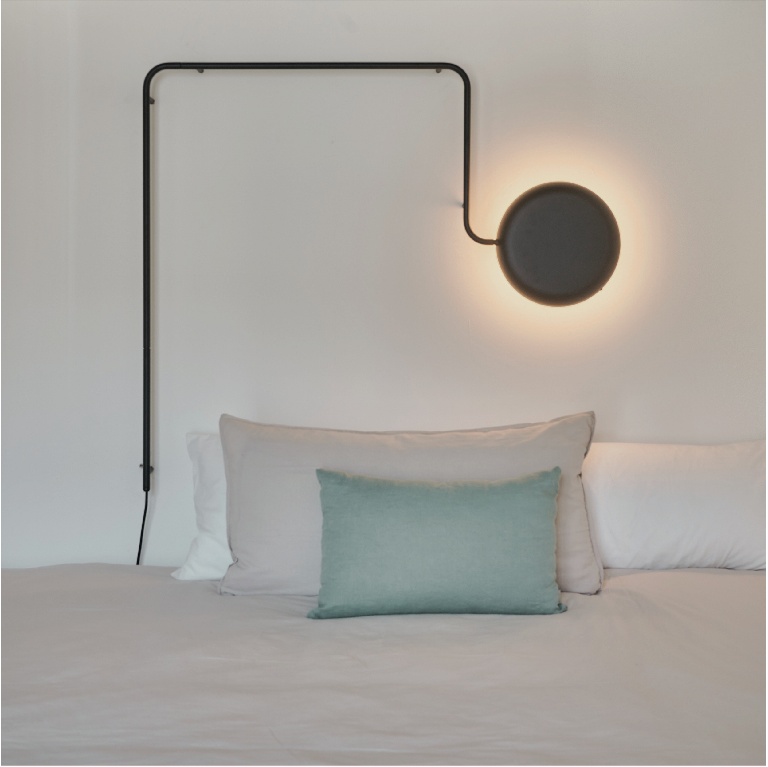 Mehrere stilvolle Kissen in Weiß, Hellblau, Grau und Beige übereinander vor einer weißen Wand, im Hintergrund eine schwarze Stehlampe mit runder Lichtquelle