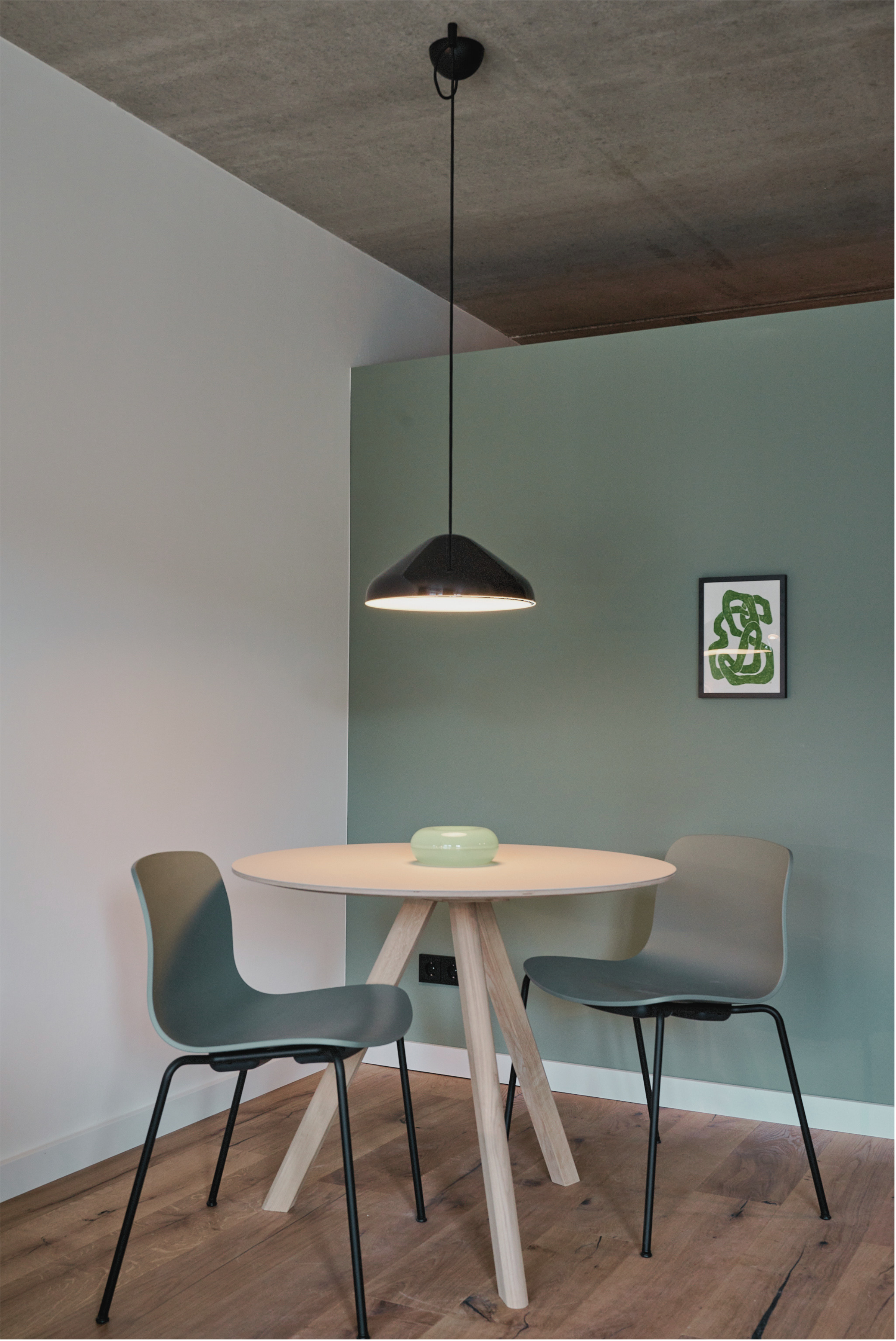 Ein runder Tisch mit drei modernen stühlen in einem Raum mit grüner und weißer Wand, hängende schwarze Lampe und ein dekoratives Bild an der Wand.