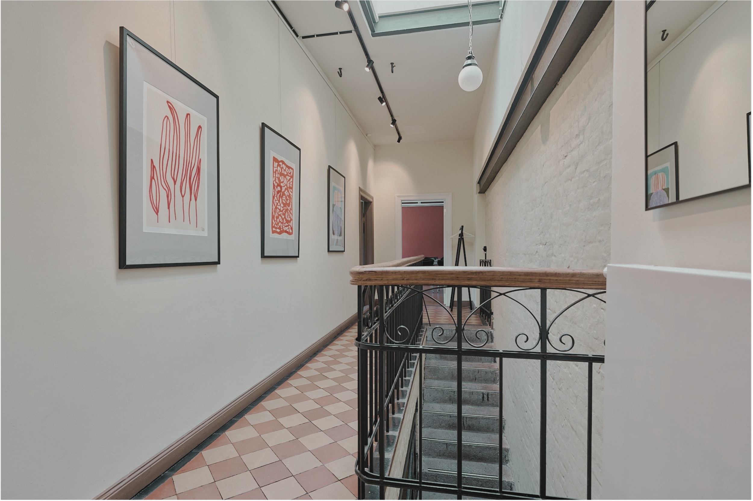 Blick auf eine Treppe in einem Kunstmuseum mit weißen Wänden, die mehrere farbenfrohe Kunstwerke in schwarzen Rahmen zeigen, und eine Deckenleuchte im oberen Bereich.