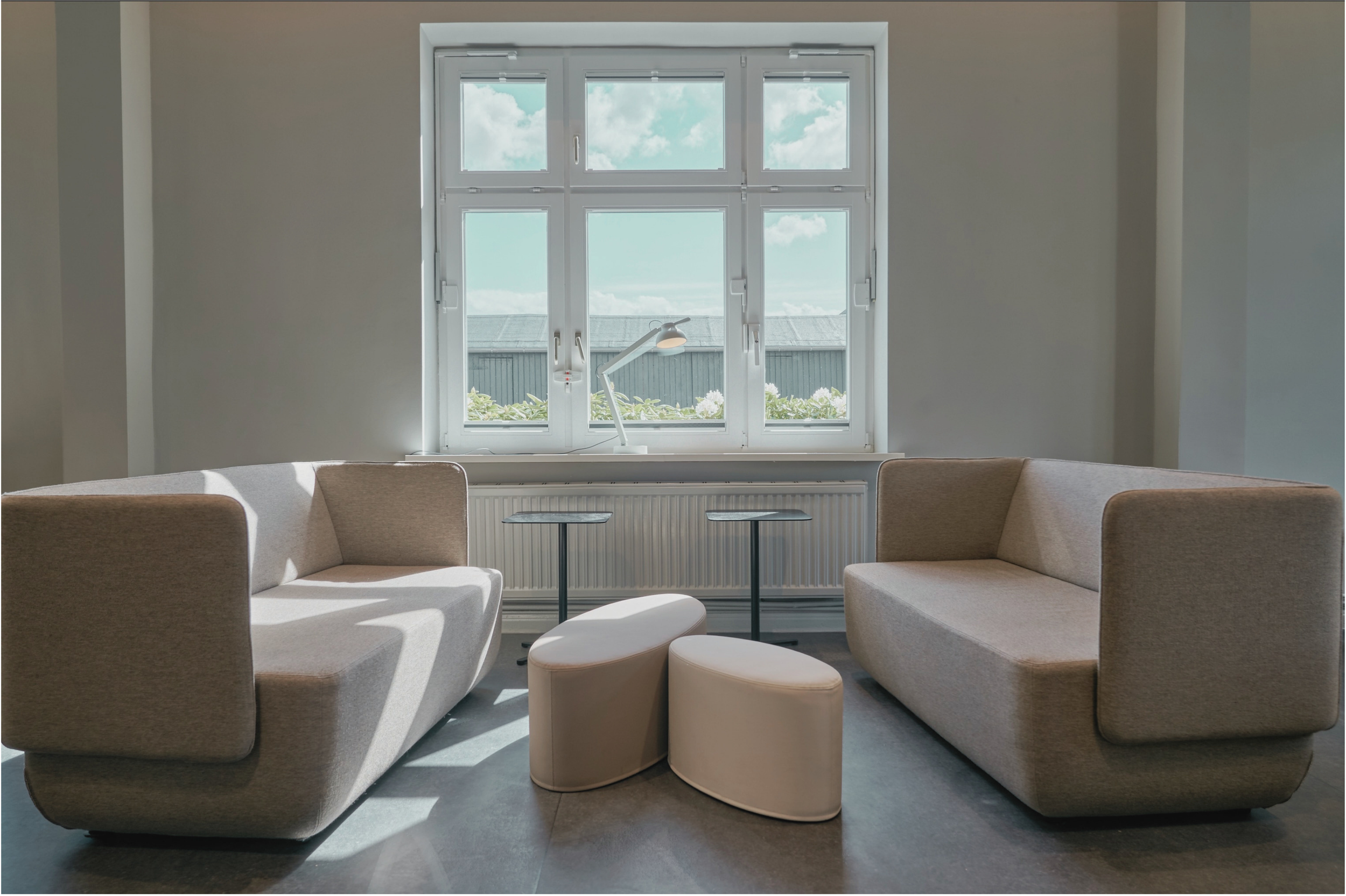 Geräumiges Wohnzimmer mit großen Fenstern, zwei beige Sofas, einem weißen Sessel und einem weißen Stehtisch, im Hintergrund eine Fensterbank mit Pflanzen und Blick auf Himmel und Dach.
