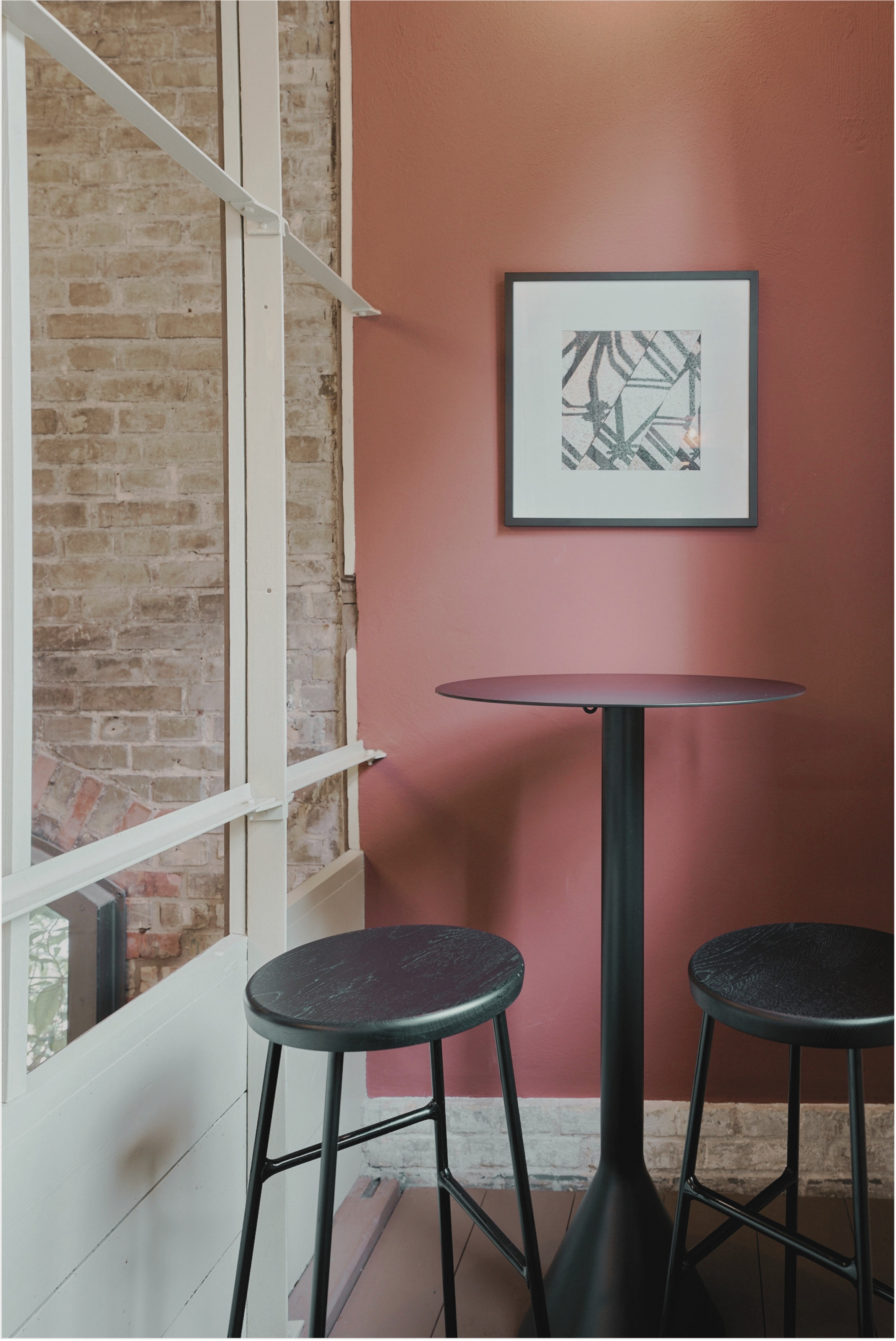 Ein gemütliches Café mit einem hohen schwarzen Tisch und zwei schwarzen Hockern vor einer roten Wand. An der Wand hängt ein abstraktes Kunstwerk in einem schwarzen Rahmen.