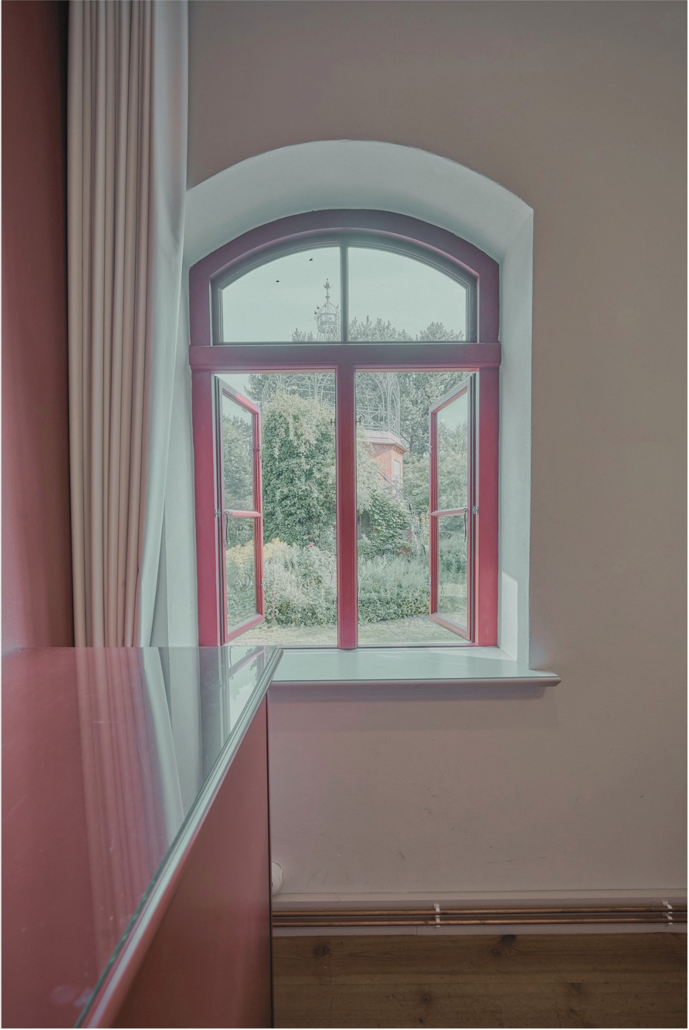 Offener Fenster mit roten Rahmen vor einer weißen Wand, Blick nach draußen auf Bäume und eine Kirche im Hintergrund, lange weiße Vorhänge auf der linken Seite, darunter eine Fensterbank.