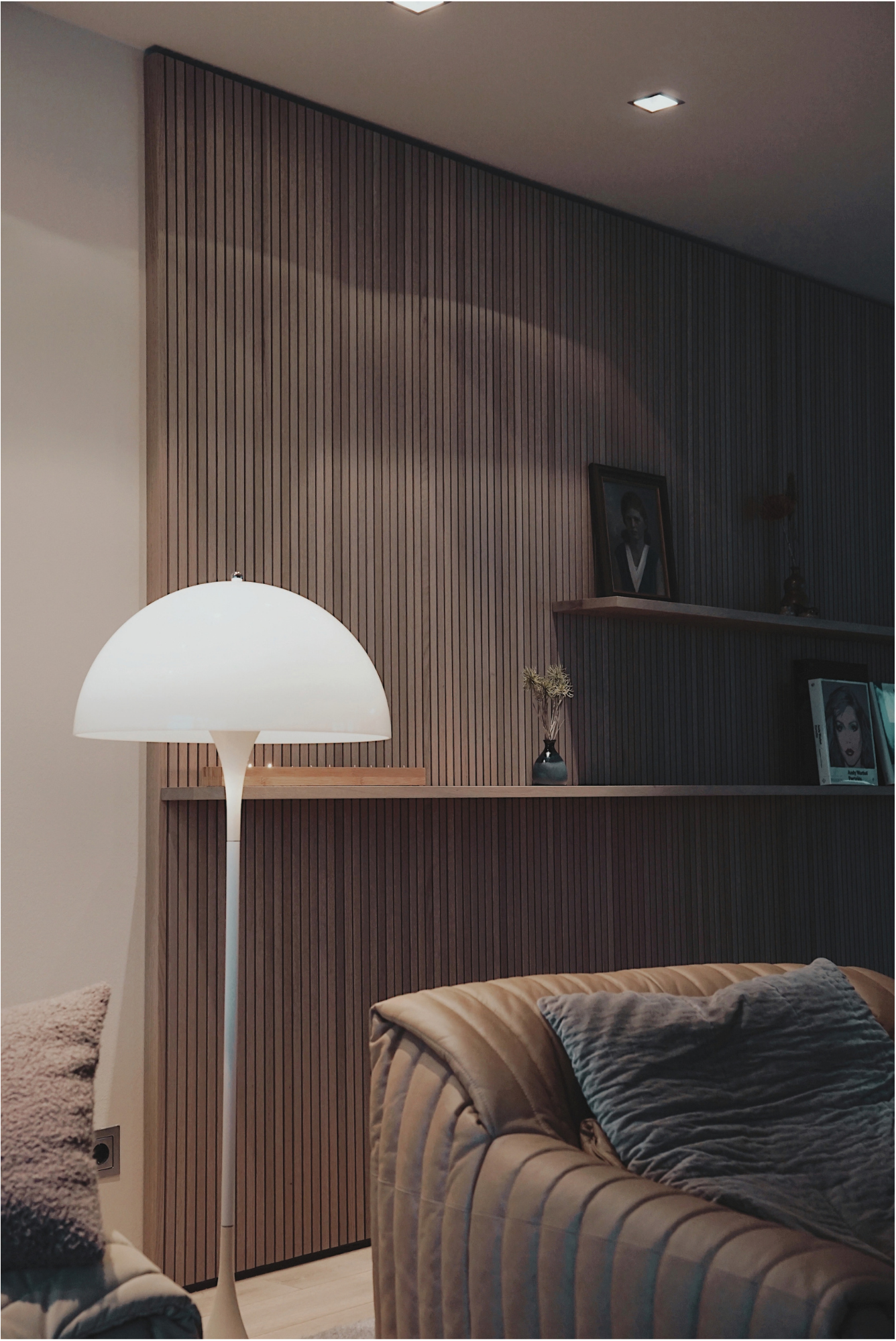 Ein Wohnzimmer mit Holzvertäfelung an der Wand, einem weißen Stehlampe, einem Bild an der Wand, einem Regal mit Dekorationen und einem Sessel mit Kissen.
