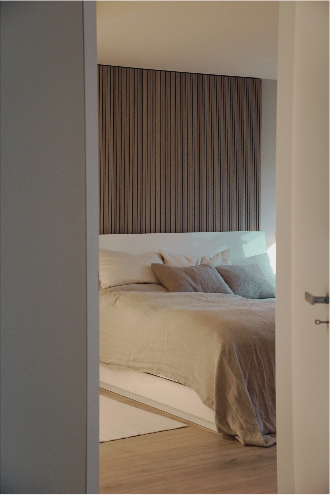 Ein Blick in ein Schlafzimmer, durch eine offene Tür. Es gibt ein Bett mit beige Bettwäsche und mehreren Kissen. Die Wand im Hintergrund hat eine Holzvertäfelung.
