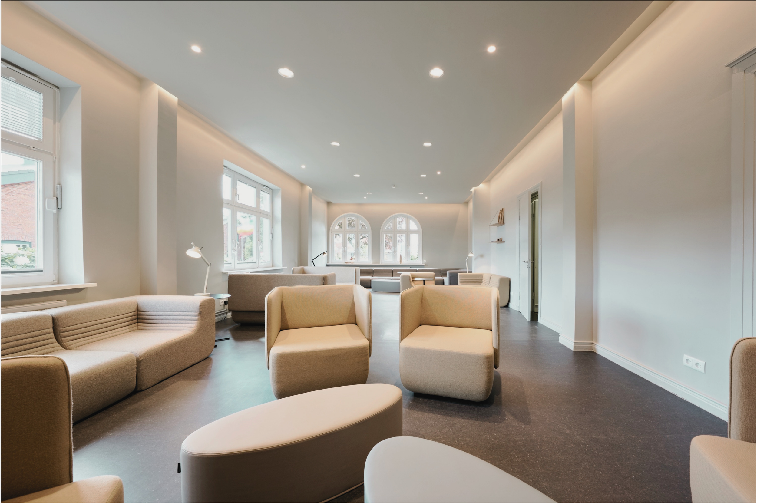 Eleganter Warte- oder Aufenthaltsraum mit bequemen beige farbigen Sesseln und weißen Tischen, große Fenster mit Blick nach draußen, helle weiße Wände, moderne Deckenbeleuchtung.