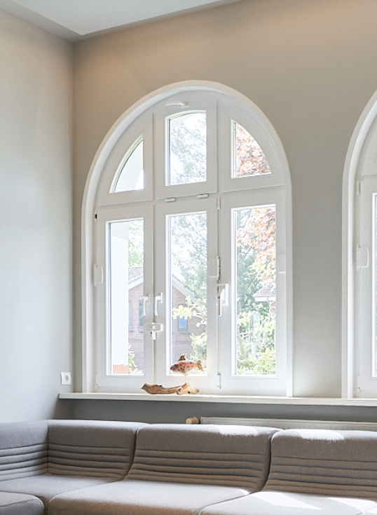 Großes, arched Fenster mit Blick nach draußen auf Bäume und Nachbarhäuser, Wohnzimmer mit grauem Sofa.