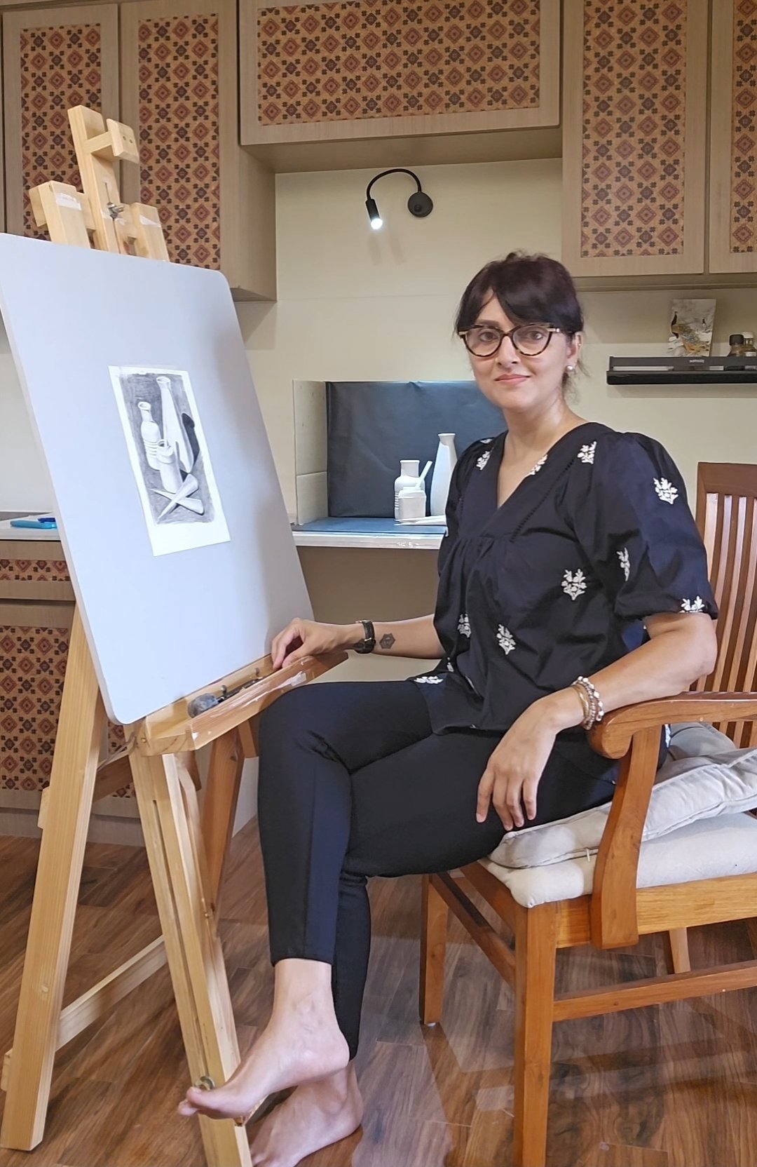 A woman with dark hair, glasses, and light skin, sitting on a wooden chair in a room, appears to be painting or drawing on a canvas on an easel, with art supplies nearby.