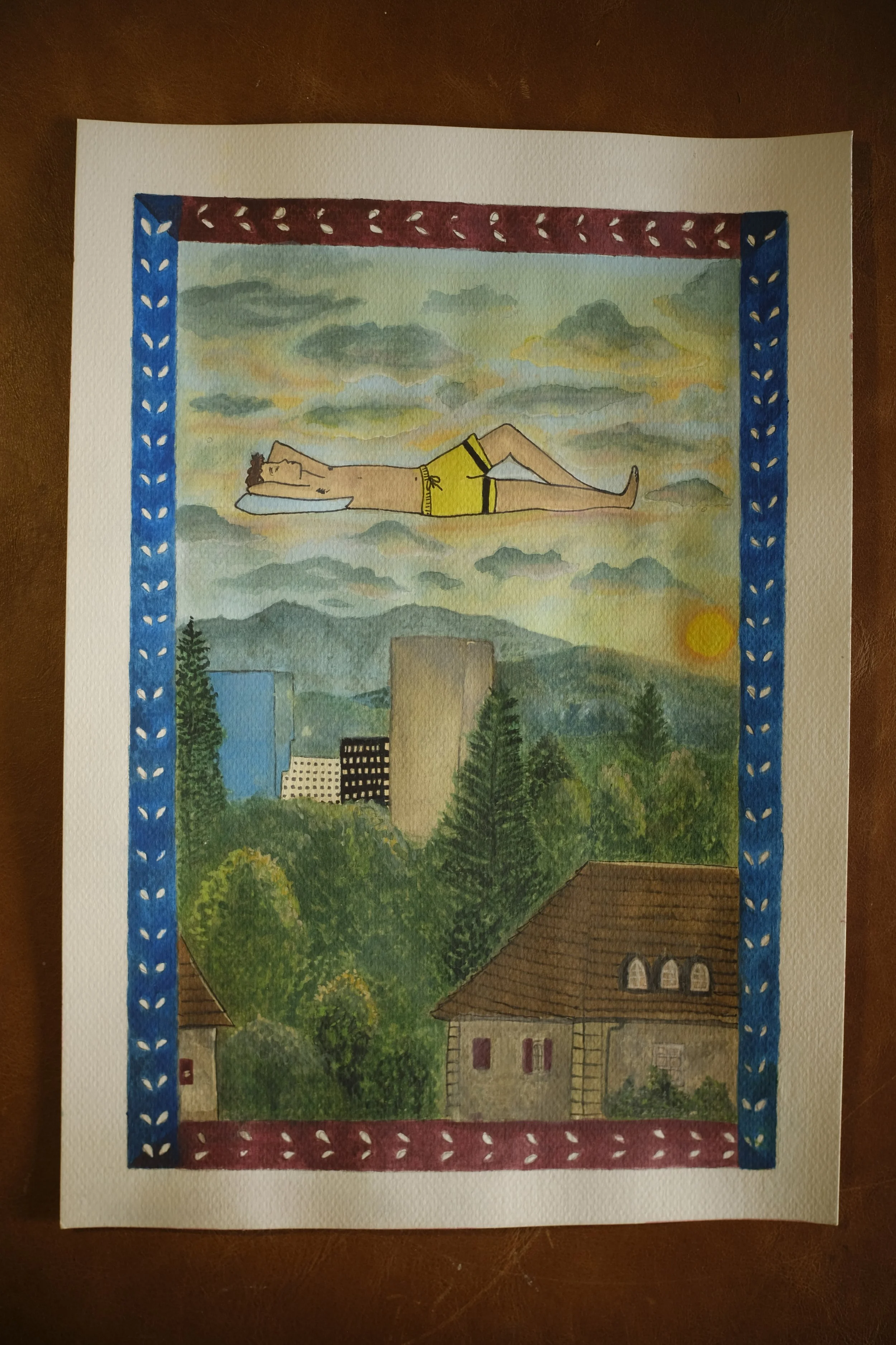 A watercolor painting on paper depicts a scene with a man lying on his back, wearing yellow shorts, with his head resting on a pillow, surrounded by clouds and a sky with the sun, framed by a decorative border of blue and red with leaf patterns.