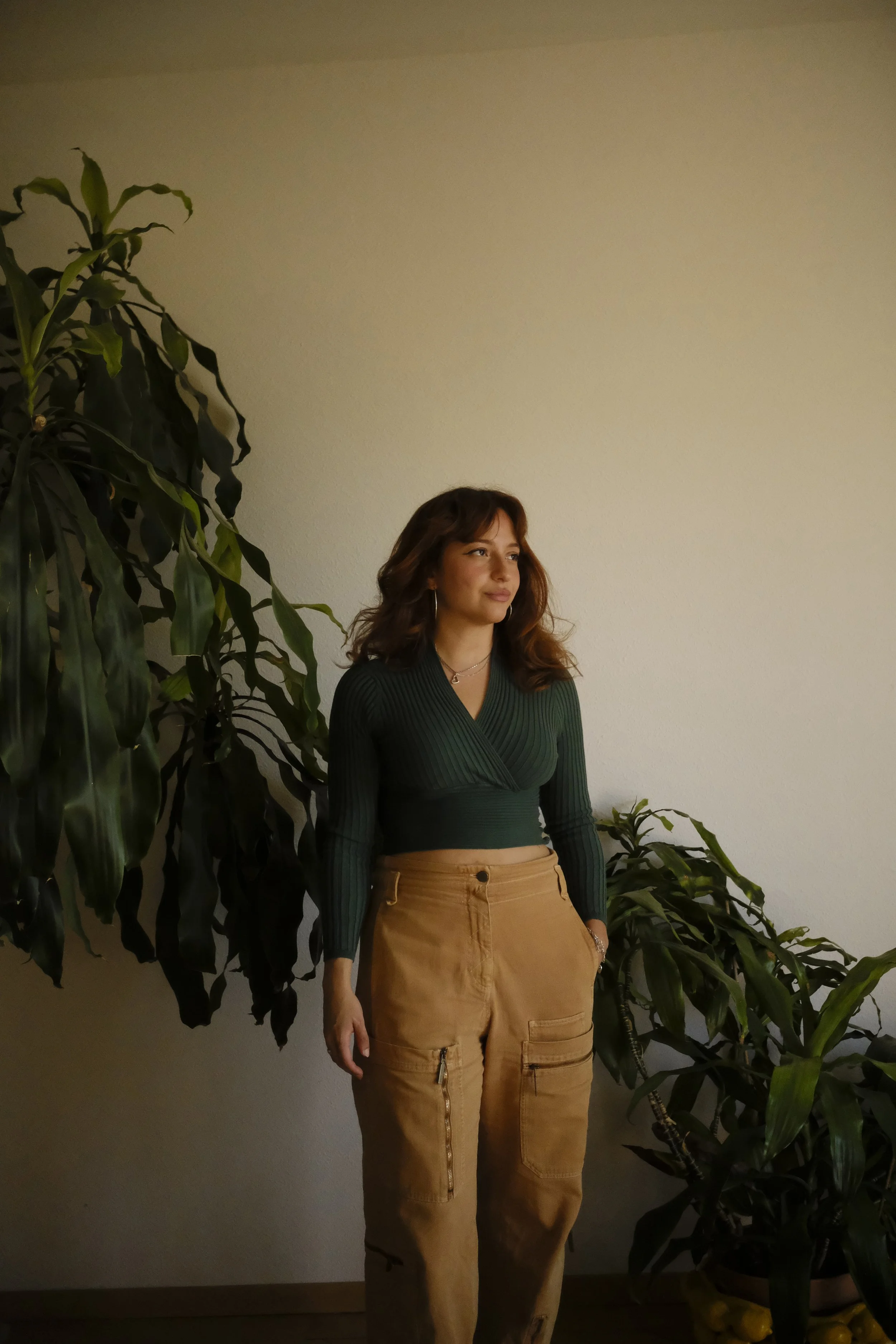 A woman with wavy brown hair wearing a dark green ribbed long-sleeve crop top and tan cargo pants standing next to large green houseplants against a plain light-colored wall.