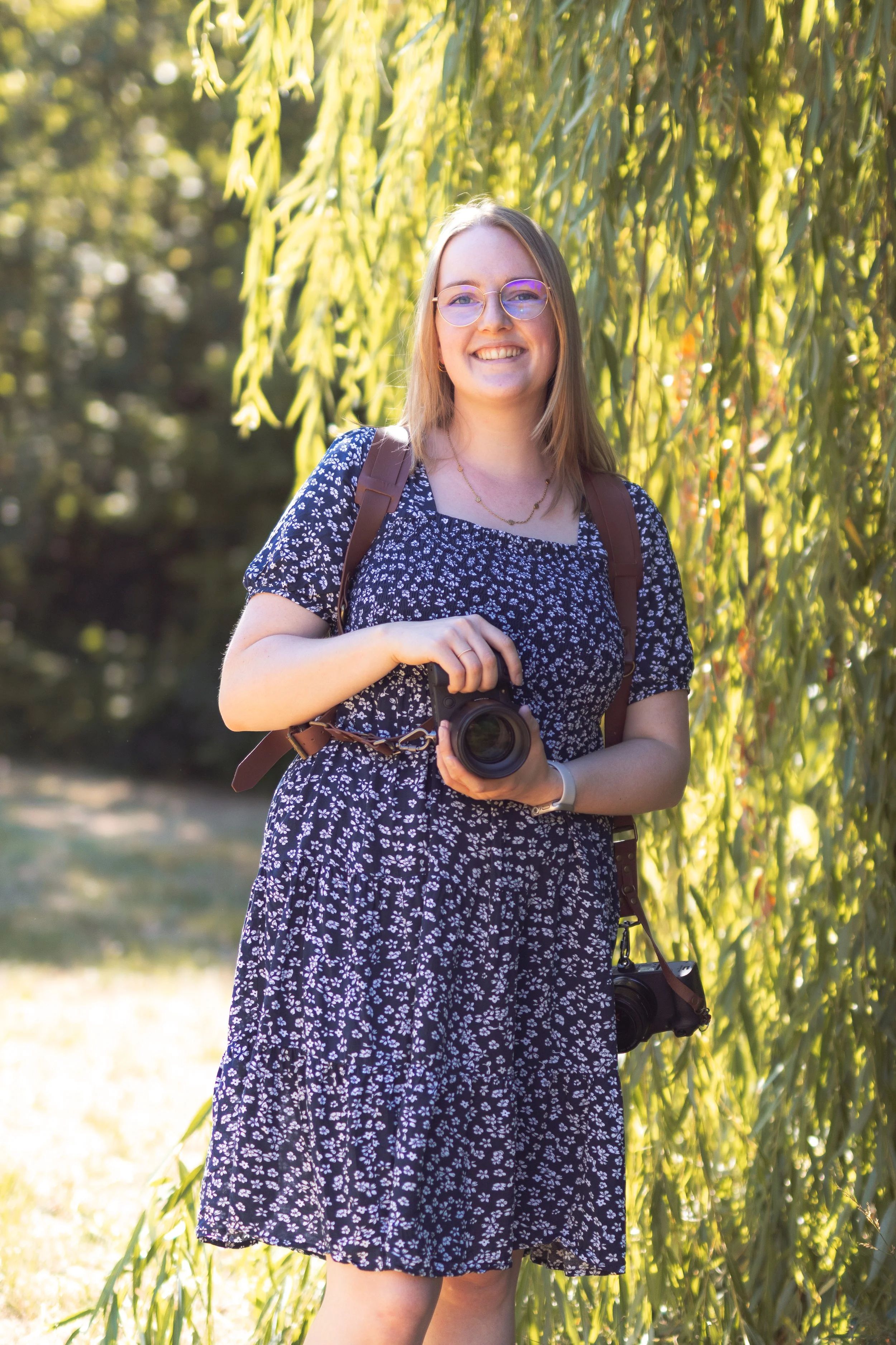 Junge Frau mit Brille, Blumenbekleidung, Rucksack und Kamera im Freien vor grünen Bäumen, sonniges Wetter.