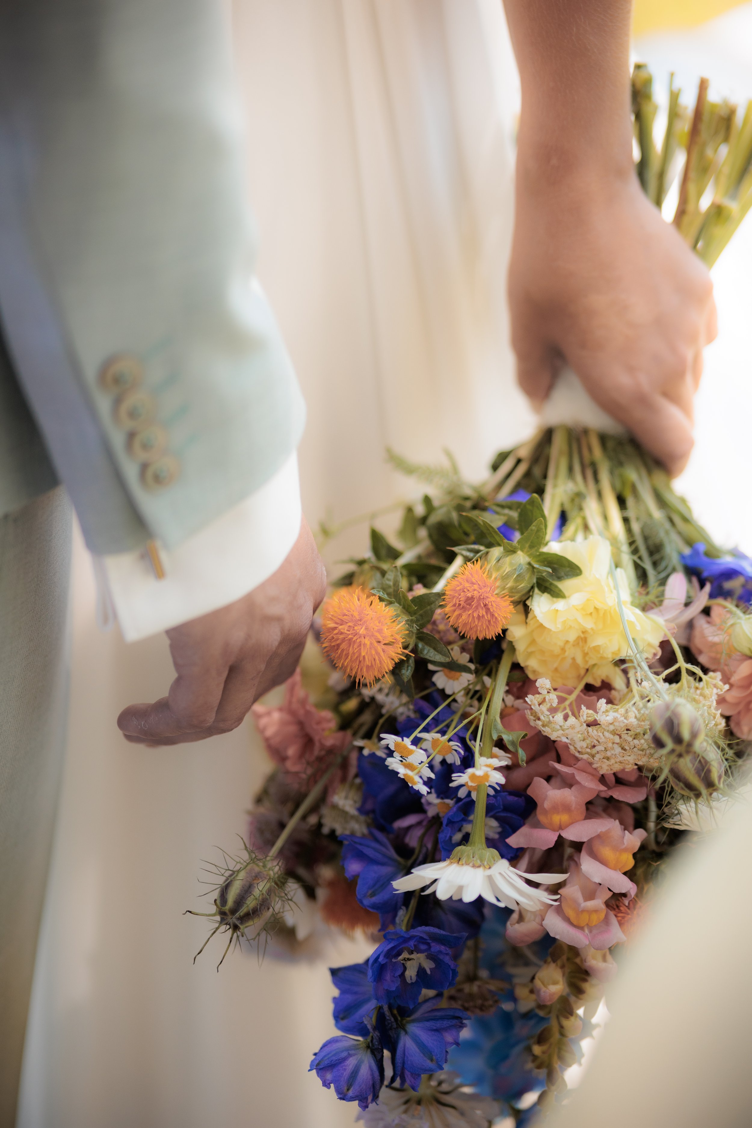 Hände, die einen Blumenstrauß halten, bestehend aus verschiedenen bunten Blumen, darunter rosa, gelbe, blaue und weiße Blüten.