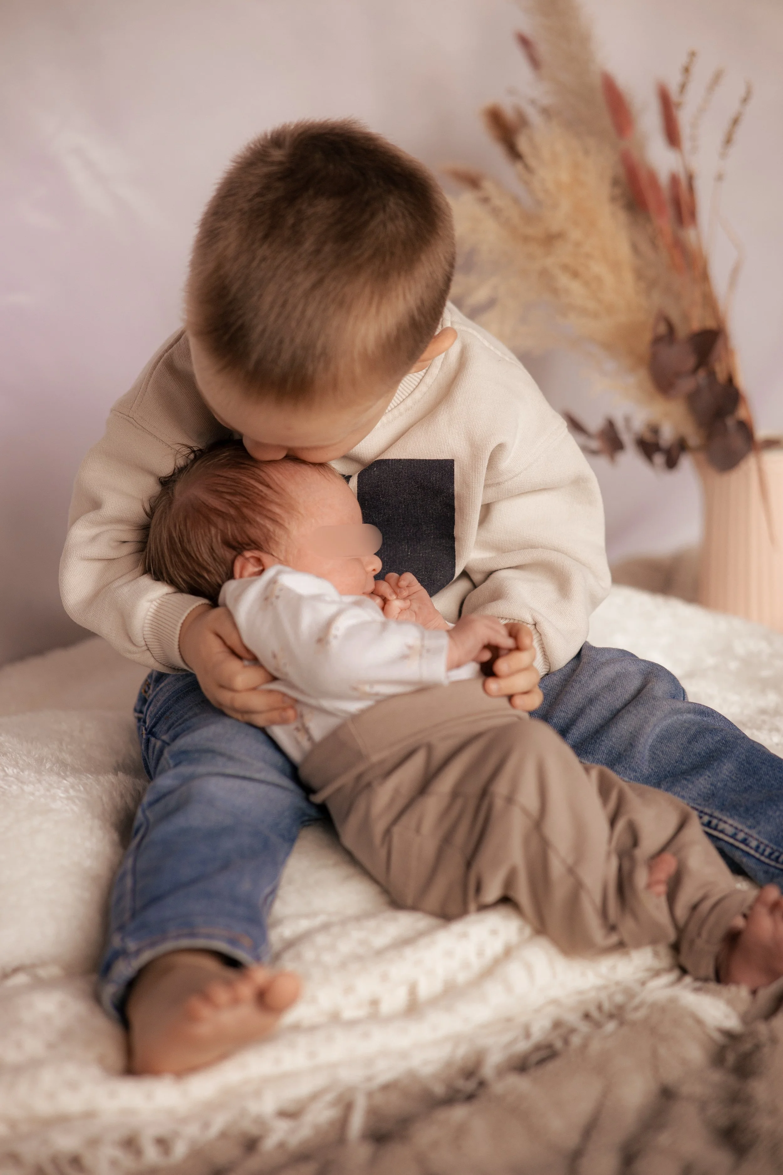 Ein älterer Bruder hält seinen Neugeborenen Geschwisterkind auf dem Schoß und küsst es auf den Kopf, während die Geschwister auf einem weißen, plüschigen Bett liegen.