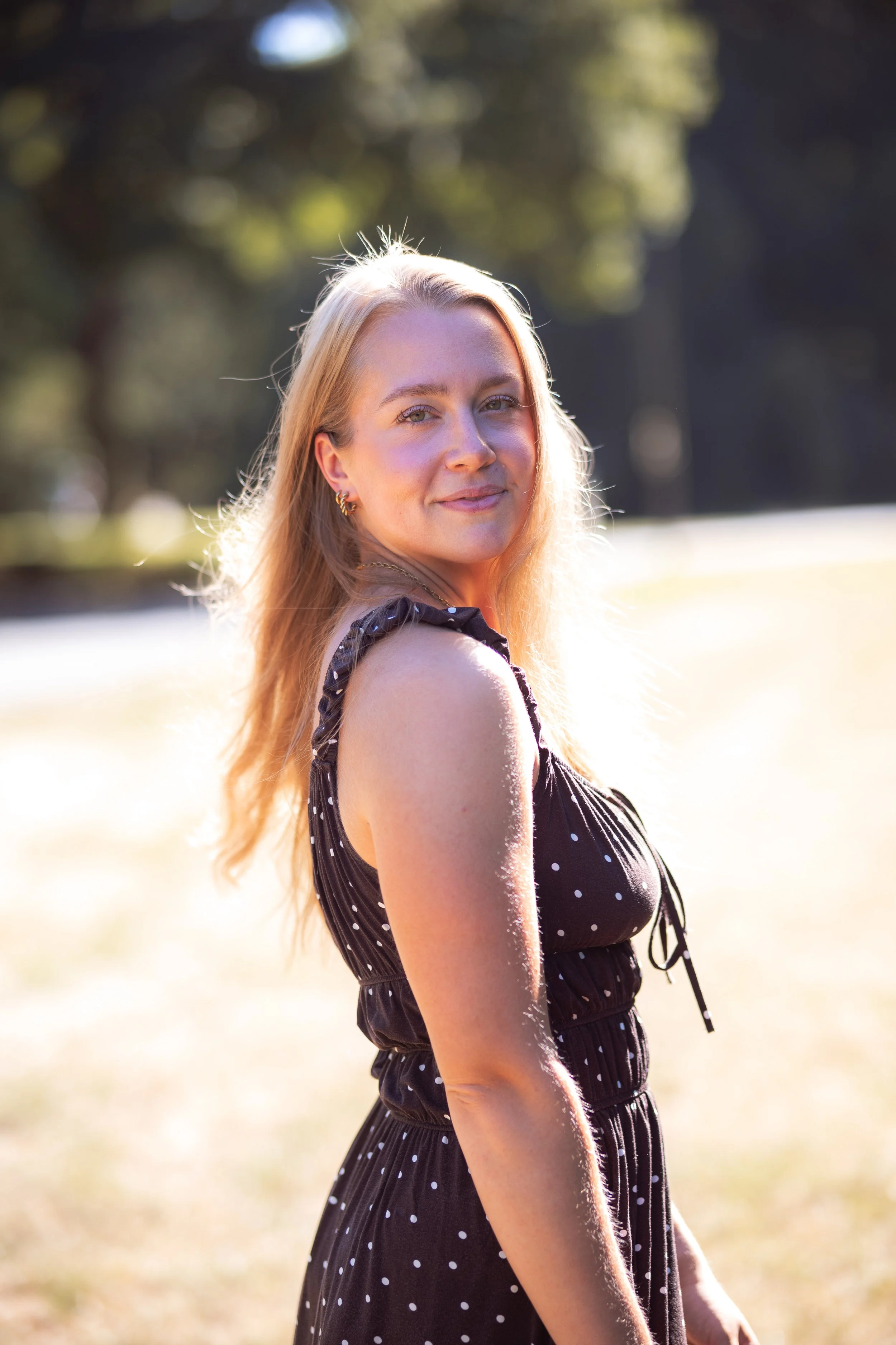 Eine junge Frau mit blonden Haaren in einem schwarzen, gepunkteten Kleid, die in der Natur steht und in die Kamera lächelt, im Hintergrund Bäume und Wasser bei Sonnenuntergang.