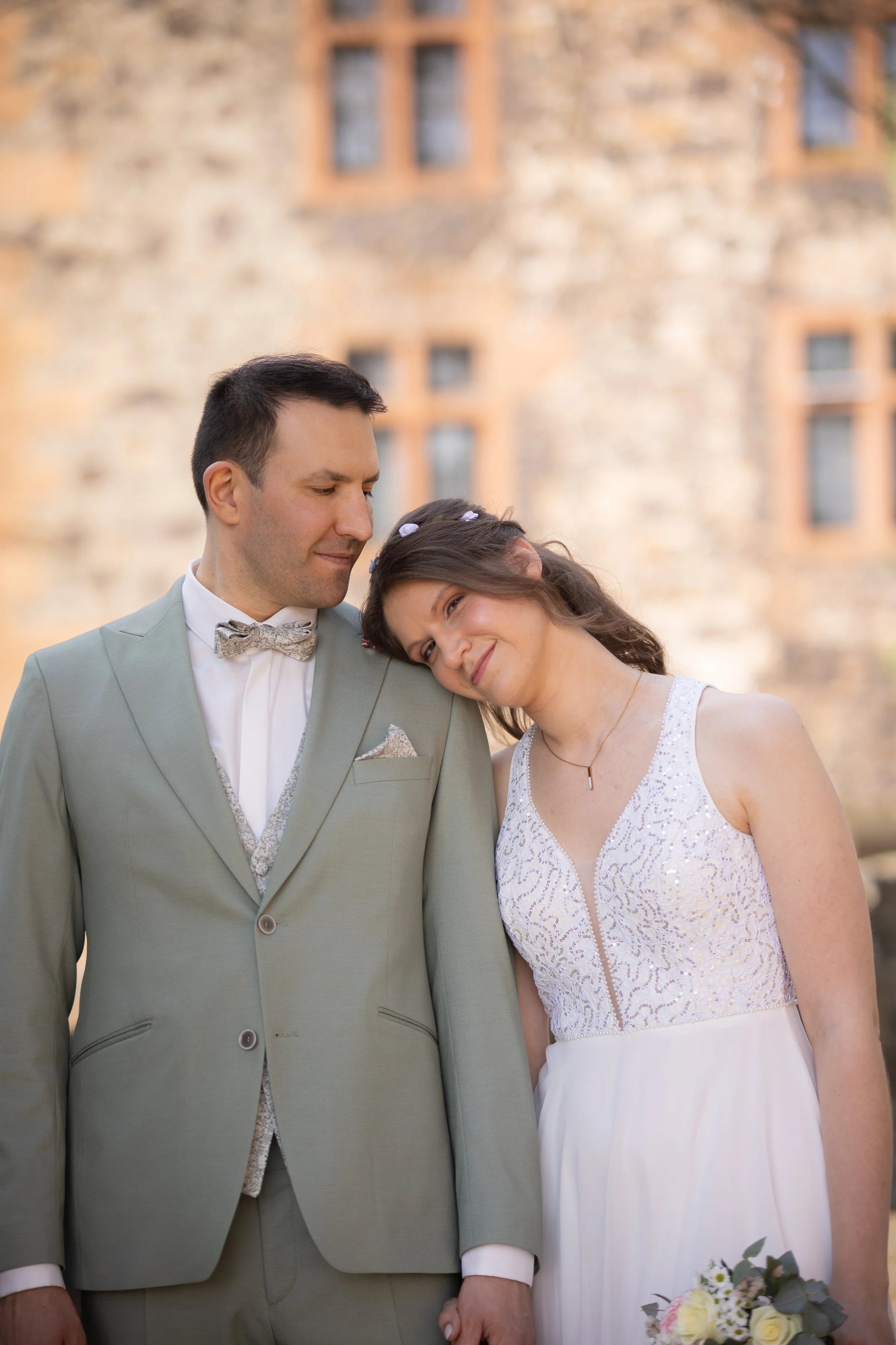 Ein Paar bei einer Hochzeit, der Mann trägt einen hellgrünen Anzug mit Fliege, die Frau trägt ein weißes Hochzeitskleid und hält einen Blumenstrauß.