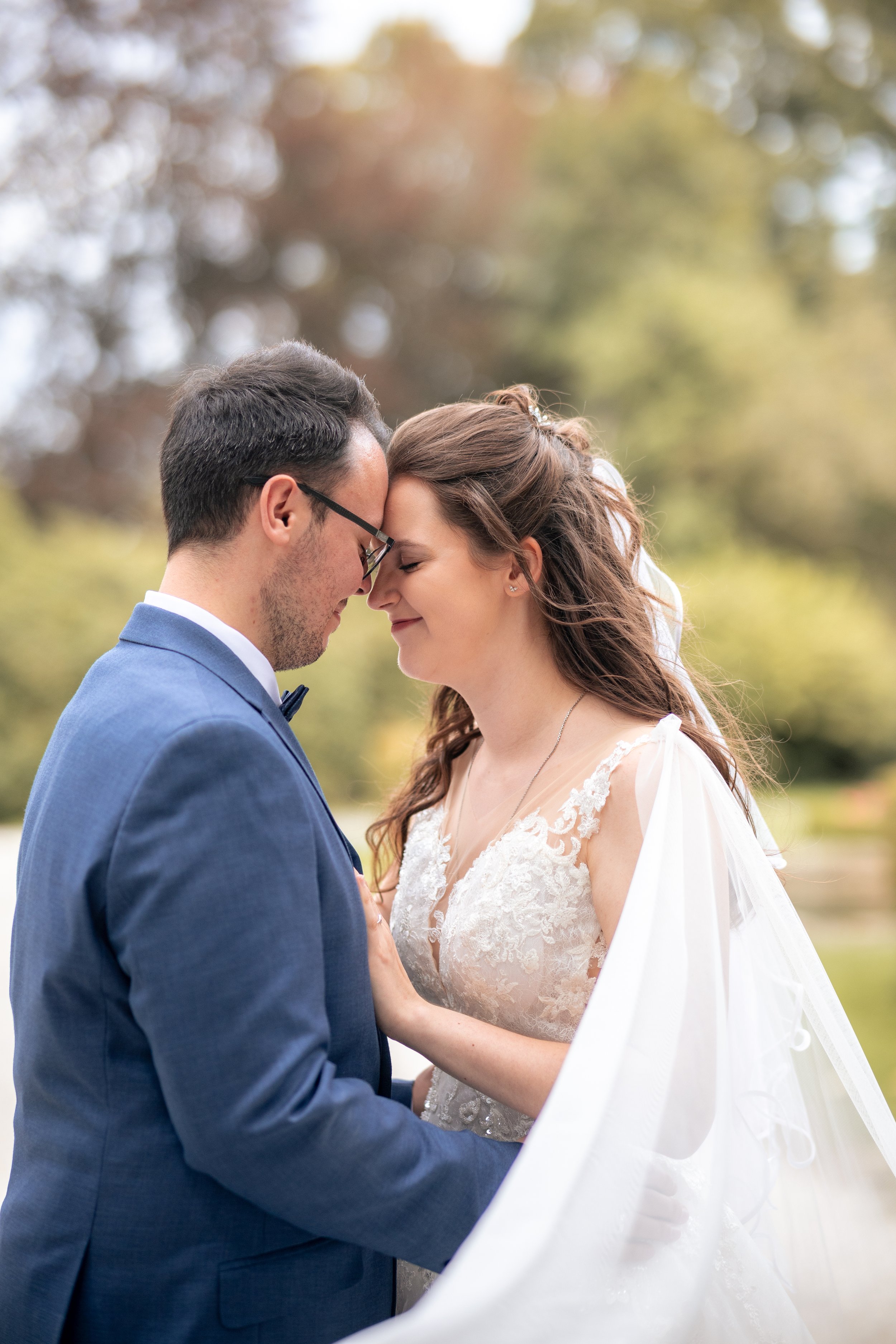Ein Brautpaar steht sich vor einem Baum im Freien sehr nahe und berührt ihre Stirnen. Es ist ein Mann in einem blauen Anzug mit Brille und eine Frau in einem weißen Brautkleid mit lockigen Haaren. Im Hintergrund sind herbstliche Bäume mit blättrigem 