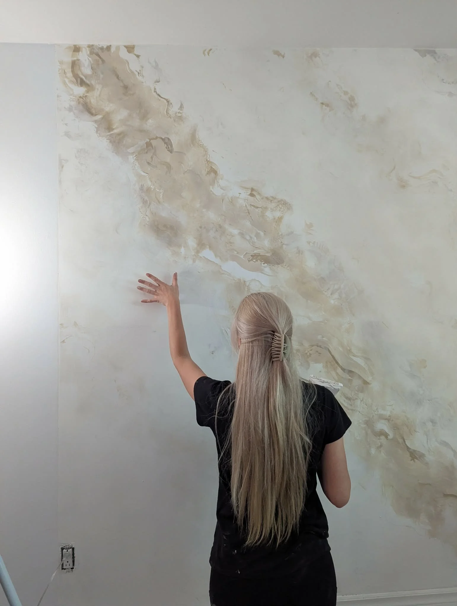 Une personne aux longs cheveux blonds portant un t-shirt noir se tient devant un mur blanc avec une texture beige, levant la main gauche.