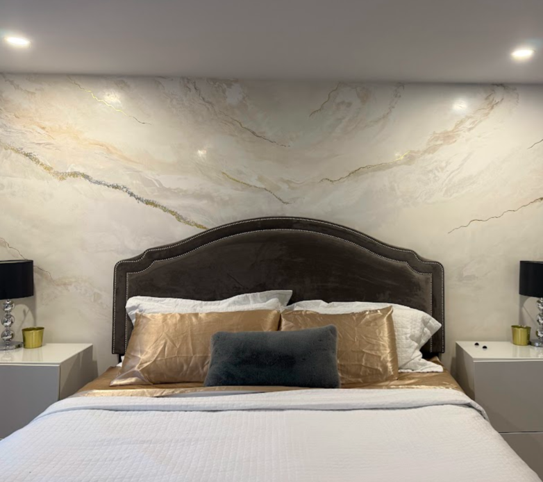 Un lit avec une tête de lit en velours gris foncé, plusieurs oreillers dont certains en satin beige et un coussin gris, flanqué de deux tables de chevet blanches avec lampes noires et décorations en verre, dans une chambre avec un mur en marbre clair