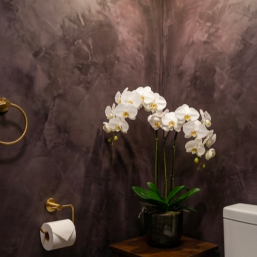 Une plante orchidée blanche dans un pot noir posée sur une table en bois, dans une salle de bain avec murs en béton et un porte-papier toilette.