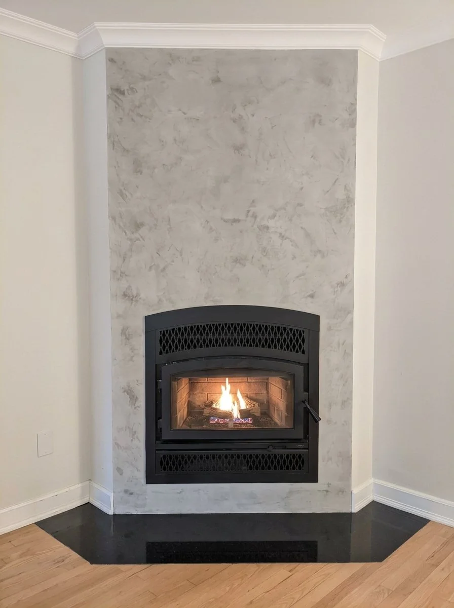 Cheminée encastrée avec feu allumé, installée dans un mur blanc, avec un revêtement en plâtre gris et un sol en bois clair.