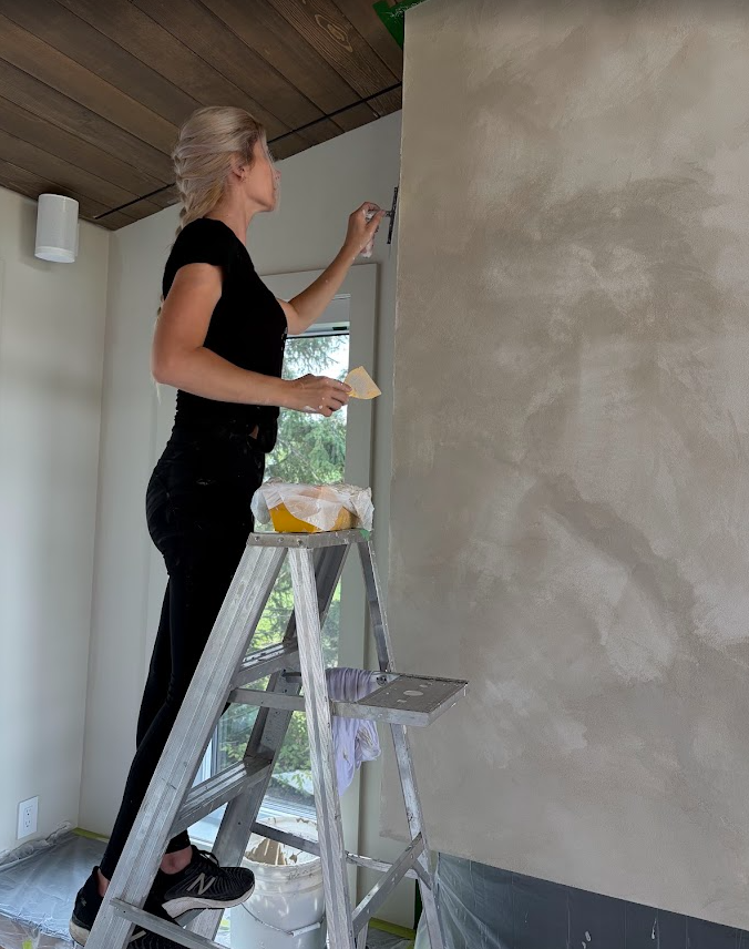 Une femme debout sur une échelle en train de peindre ou de faire des retouches sur un mur intérieur beige avec un rouleau ou un pinceau, dans une pièce lumineuse avec un plafond en bois.