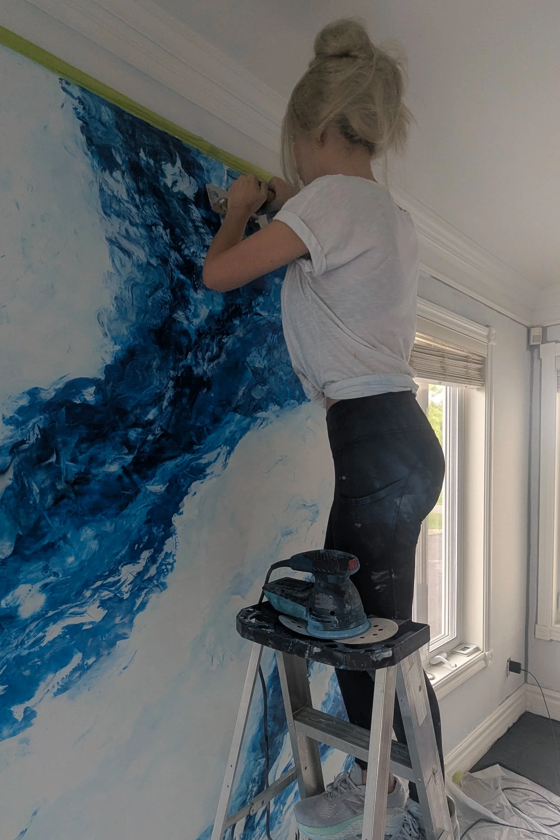 Une femme peignant une murale abstraite avec des tons bleus, la femme est sur une échelle, portant un t-shirt blanc et des pantalons noirs, le sol est recouvert de protection.