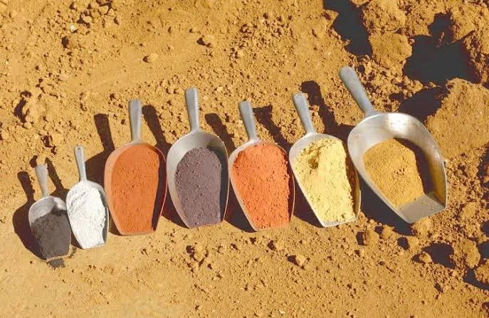 Ensemble de six moules en métal contenant différentes poudres colorées, disposés sur un sol sablonneux.