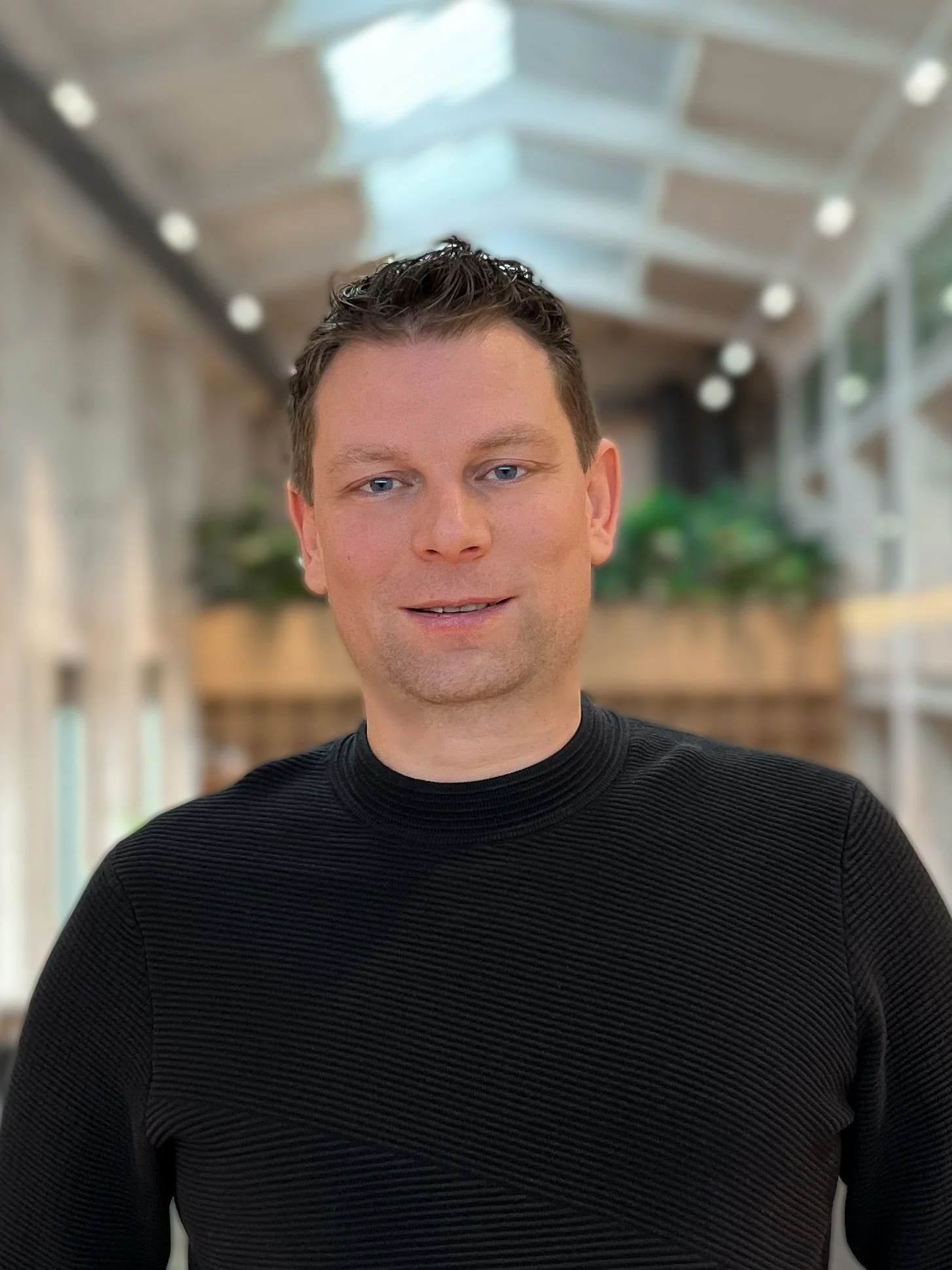 Portret van een jonge man met zwart haar, blauwe ogen en een lichte huid, gekleed in een zwart getailleerd shirt, binnen een modern gebouw met een glazen plafond en groene planten.