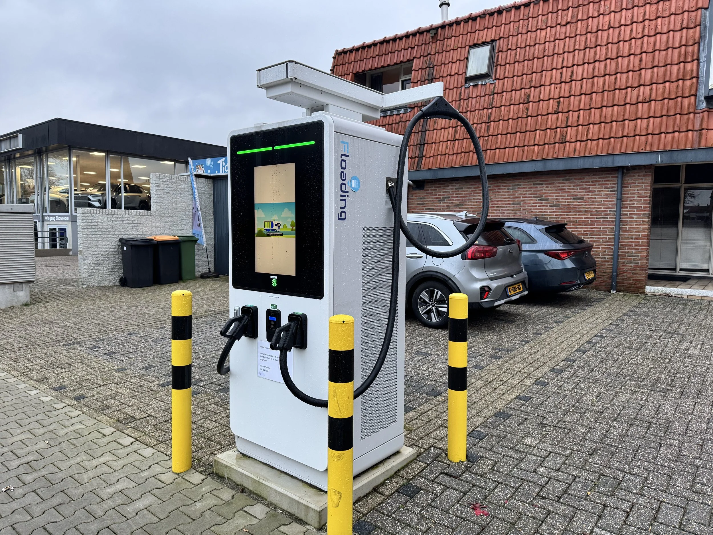 Een elektrisch laadstation voor auto's met een kabel en stekker, omgeven door gele en zwarte afzetpalen, voor een parkeerplaats met auto's en gebouwen op de achtergrond.