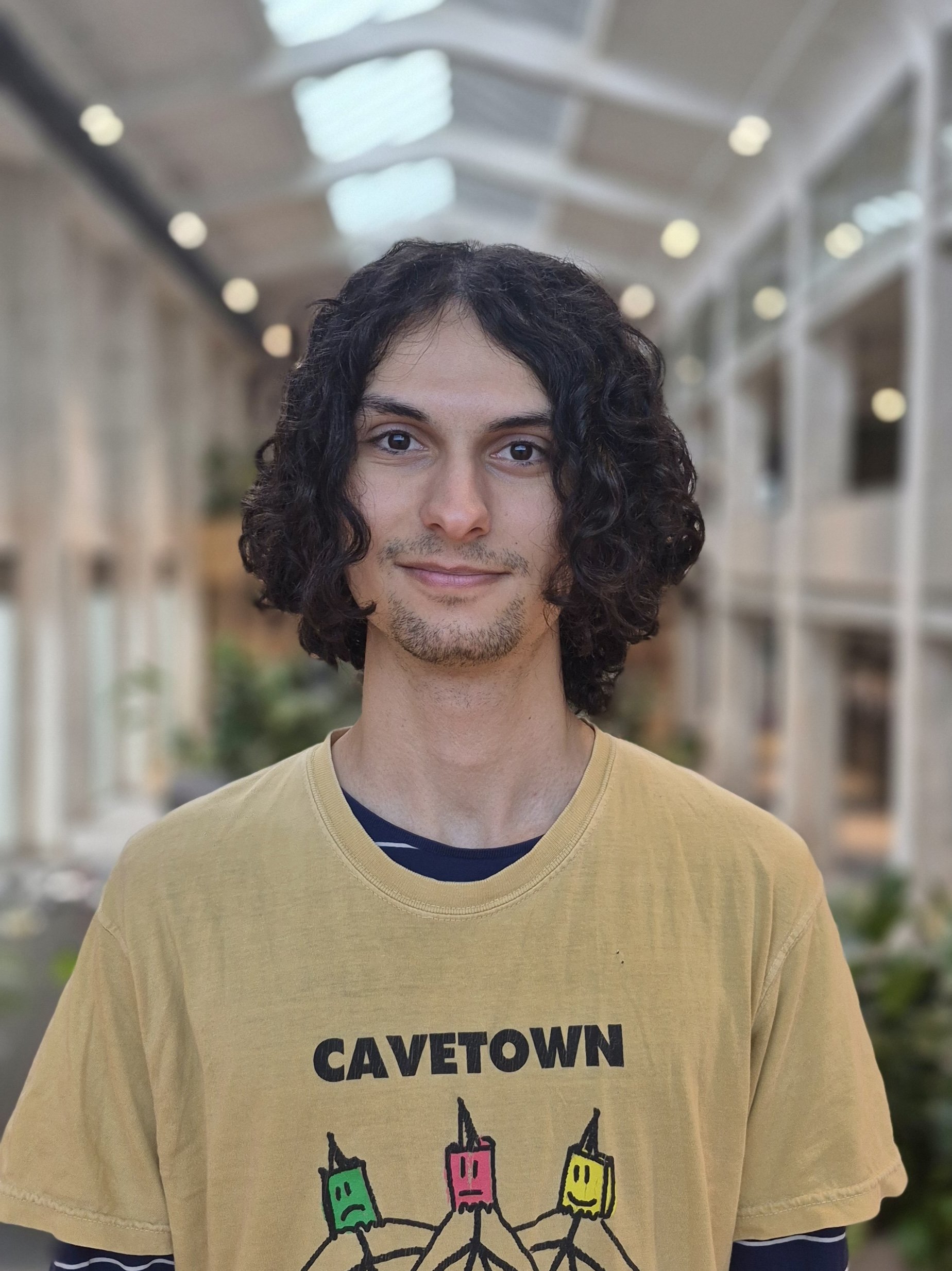 een jonge man met donker, krullend haar en lichte huid, lachend, draag een gele T-shirt met de tekst 'CAVETOWN' en een tekening van kleurrijke appel- en banaantjes vastgehouden door handen in een overdekte winkelstraat of arcade.