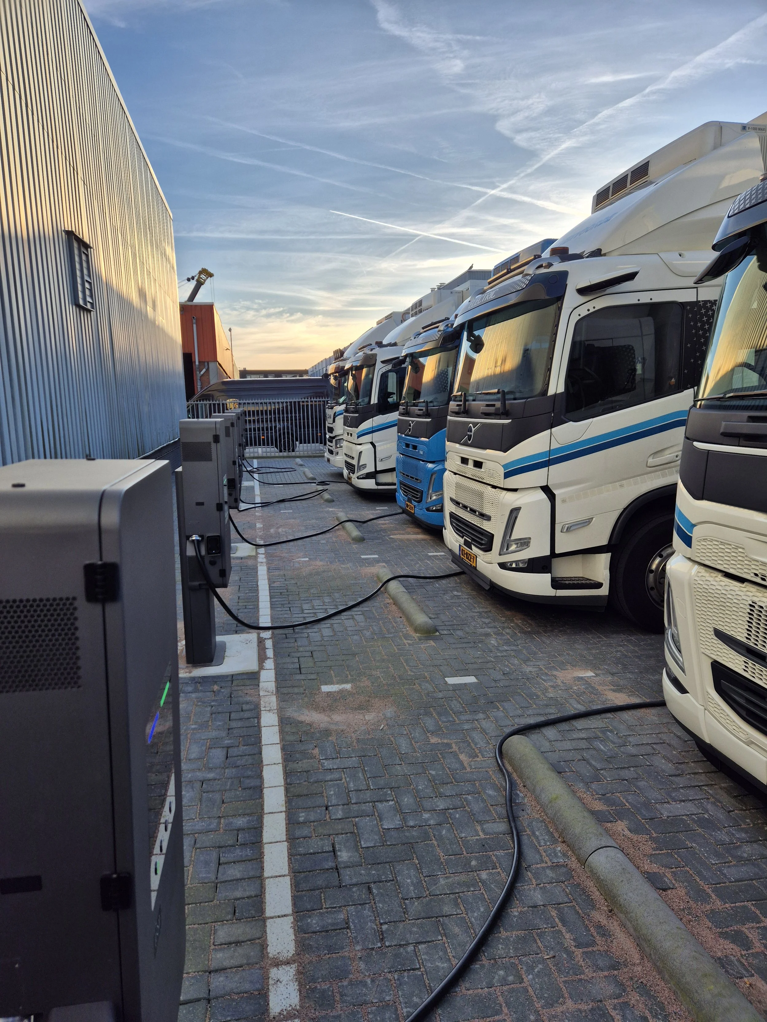 Rijen elektrische vrachtwagens opladen buiten een magazijn of logistiek centrum bij zonsondergang.