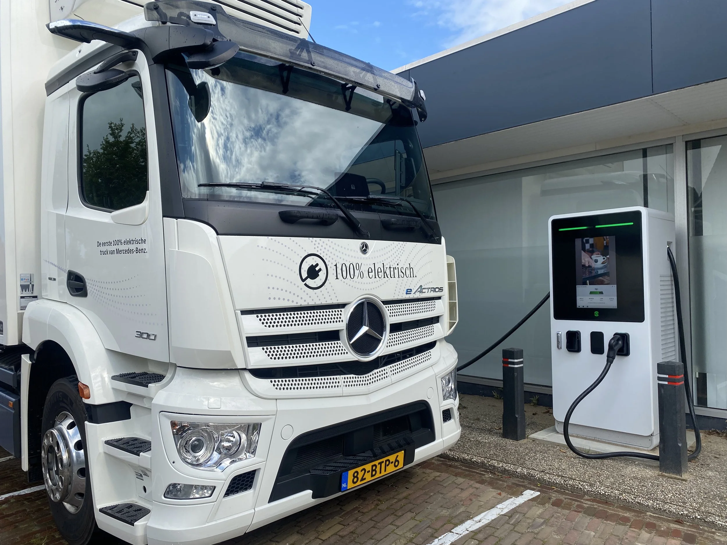 Een witte vrachtwagen die elektrisch wordt geladen bij een laadstation met een tijddisplay en laadkabel.