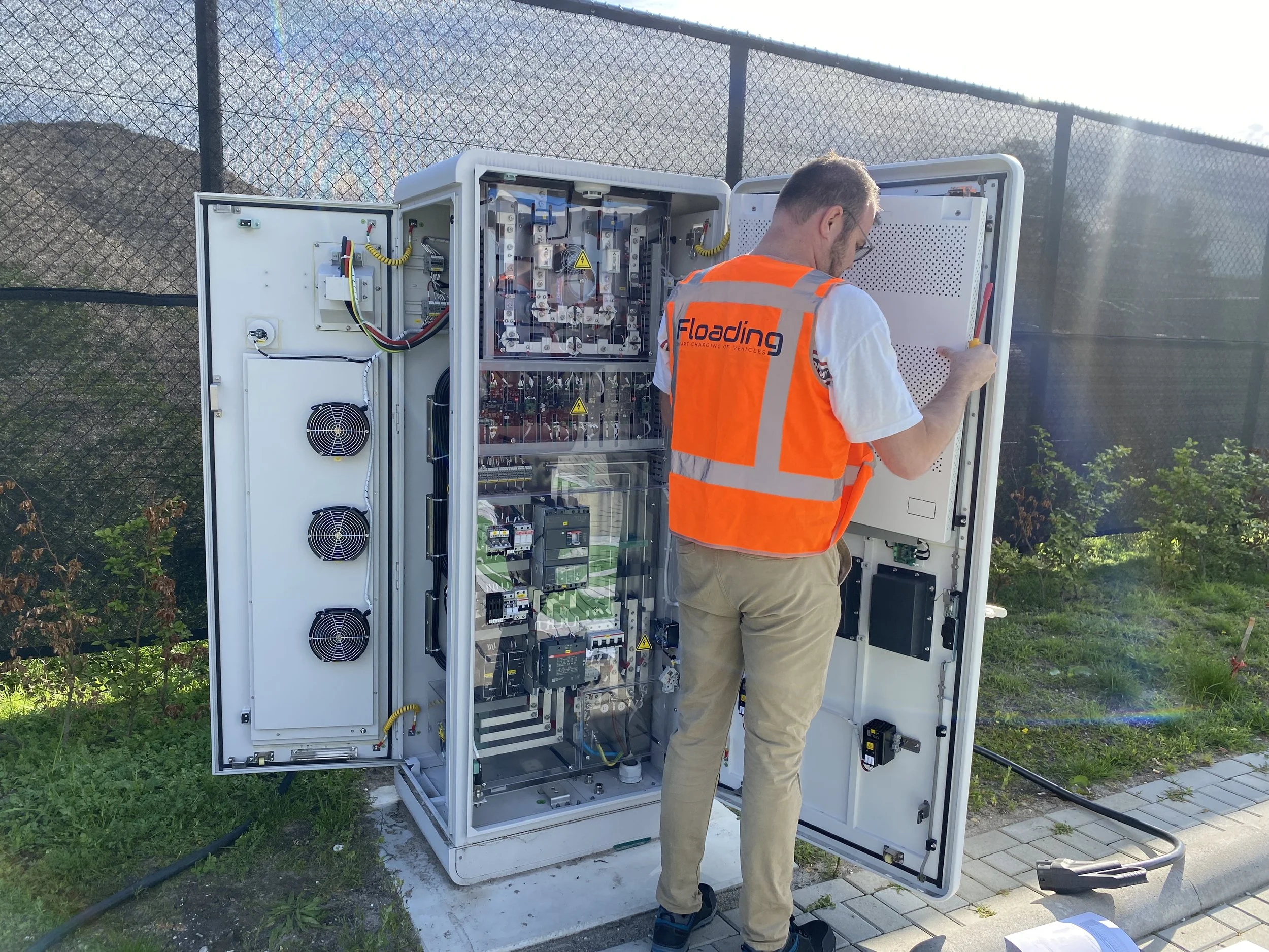 Man onderzoekt een open elektrisch bedieningskast met interne elektrische apparatuur, buiten naast een hek en groene planten.
