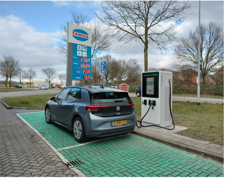 Een elektrische auto wordt opgeladen bij een tankstation op een elektrische laadpaal. Op de achtergrond is een scherm te zien met de gasprijzen.