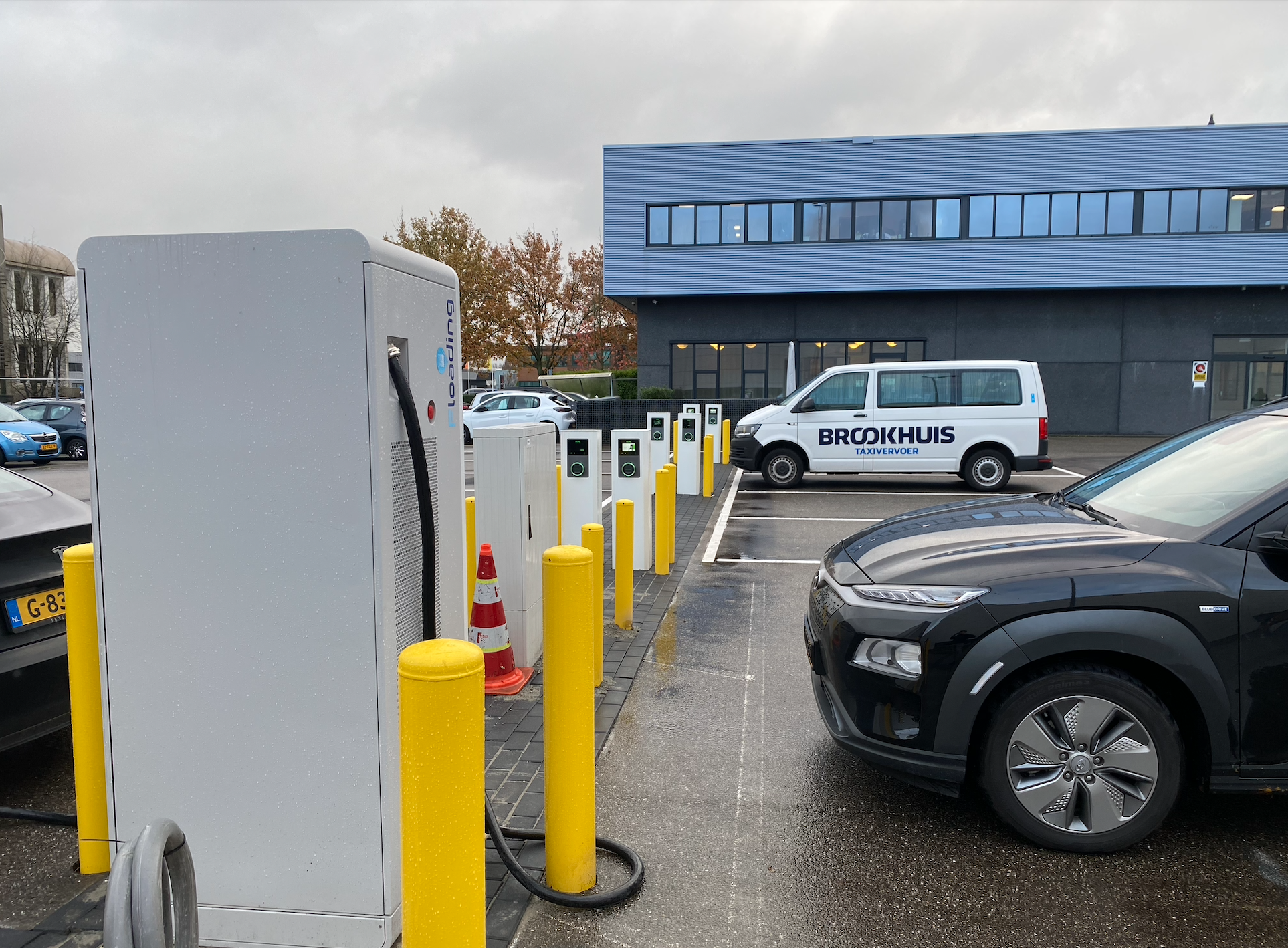 Elektrische laadpaal voor een auto, parkeerplaats met een busje met de tekst 'Brokhuis Taxivervoer', en een modern gebouw op de achtergrond.