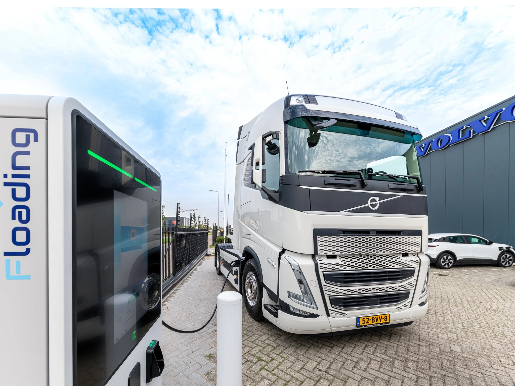 Een witte vrachtwagen die wordt opgeladen bij een elektrische laadpaal, met een kade en bedrijfspand op de achtergrond onder een bewolkte hemel.