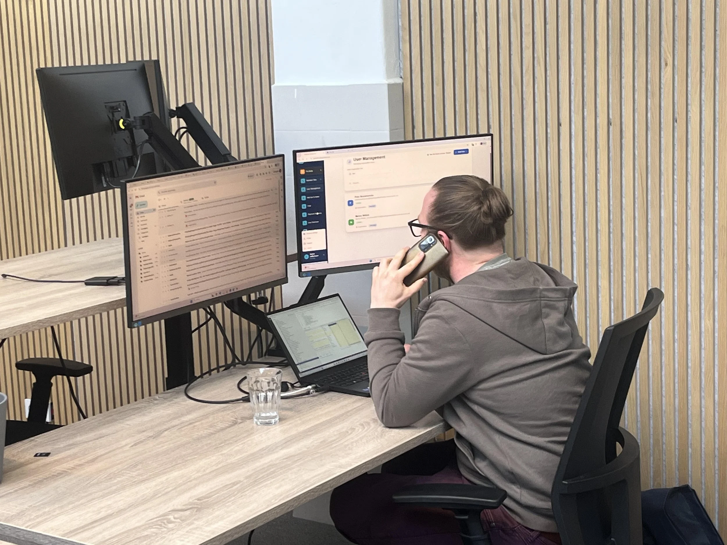 Man zit aan een houten tafel met twee computerschermen, één gebruikt en één uit, en belt met zijn mobiele telefoon, in een kantoor met houten wandpanelen.