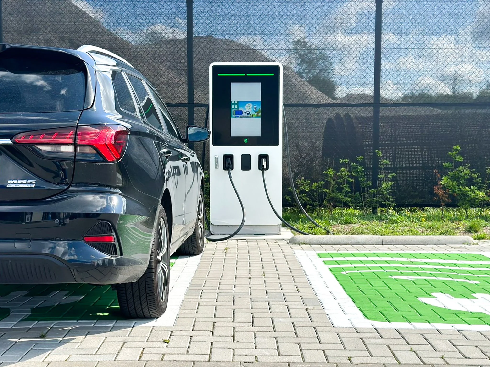 Elektrische laadpaal naast een auto op een parkeerplaats, met een groene afdrukmat op de grond.