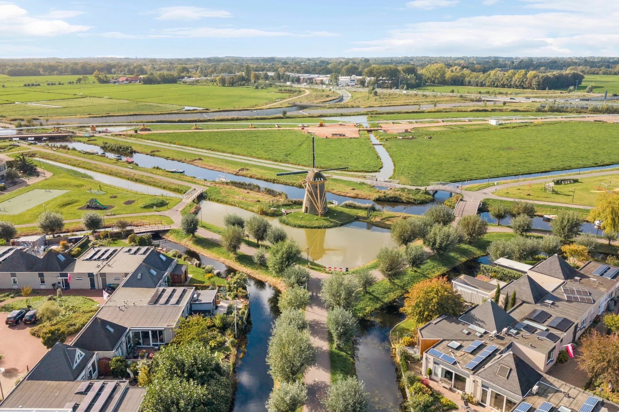 Luchtfoto van een landelijk gebied met weilanden, waterwegen, wegwerken en een woonwijk op de voorgrond. 