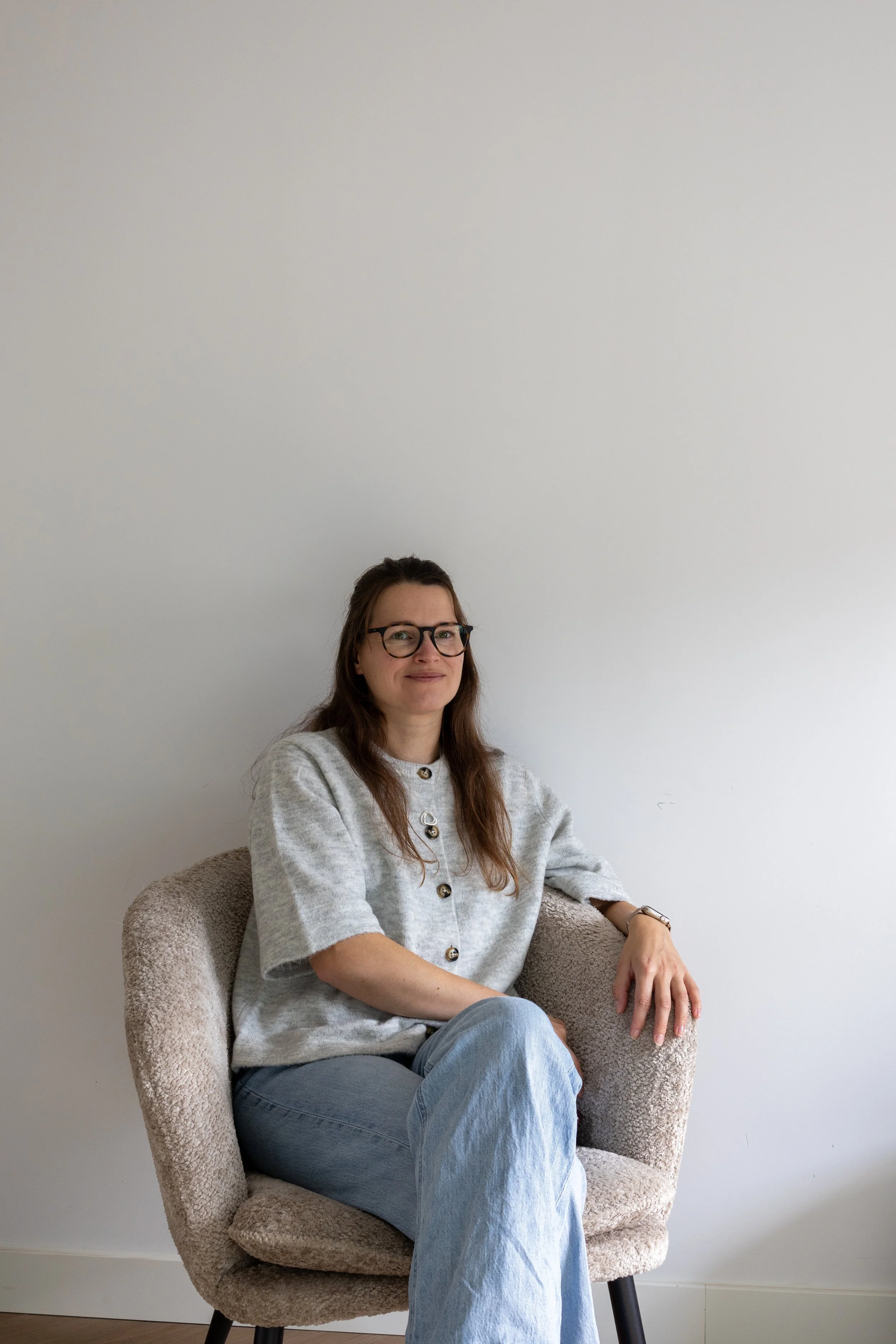Vrouw met bruin haar, brillen, grijs vest en jeans zit ontspannen in een beige stoel voor een witte muur.