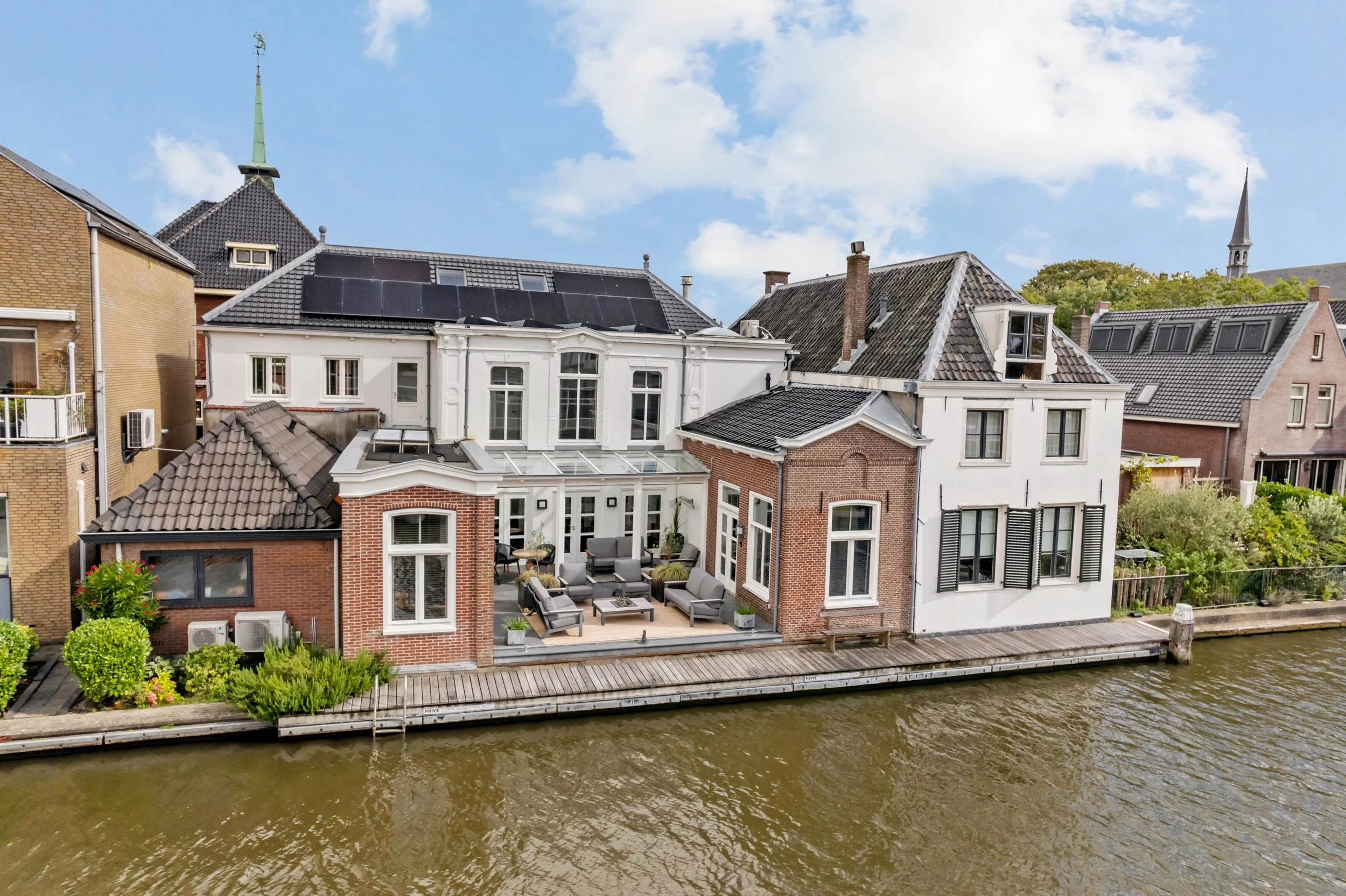 De afbeelding toont een rij huizen langs een rivier, met op de voorgrond een terras met zitmeubilair en planten. De huizen hebben verschillende dakbedekkingen, inclusief zonnepanelen en ramen.