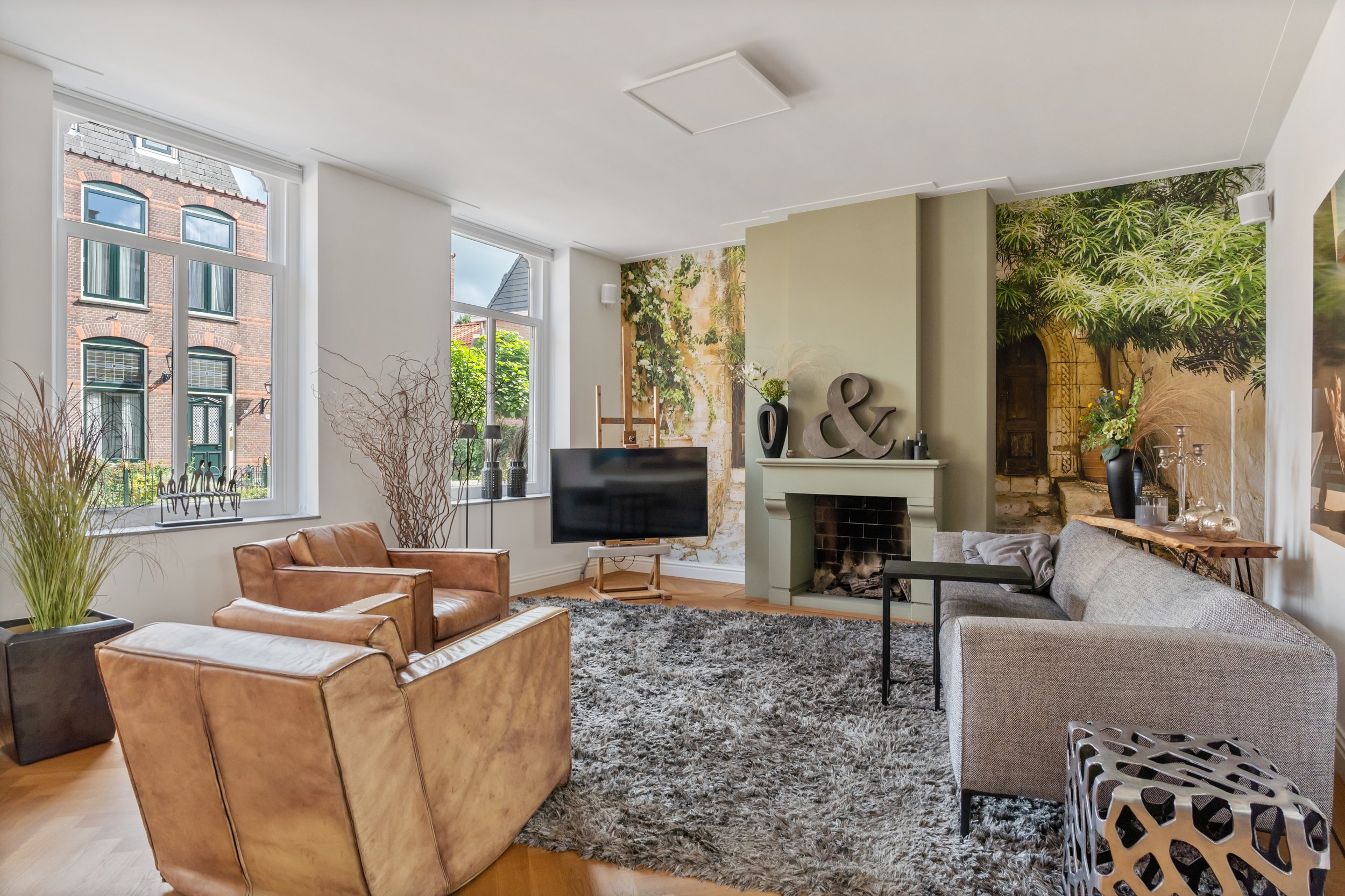 Woonkamer met grote ramen, bank, leren fauteuils, televisie, open haard, muurdecoraties en planten.