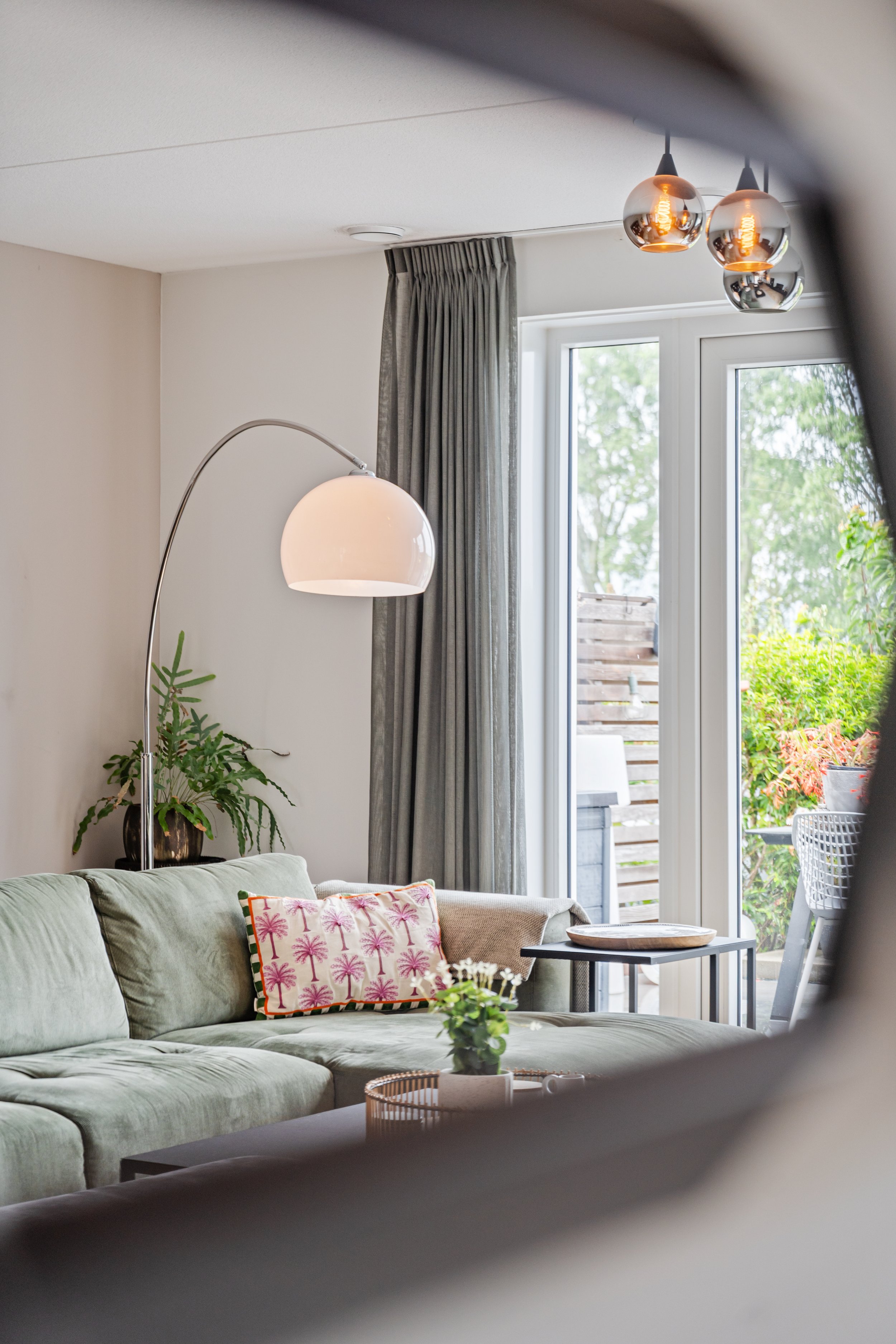 Gezellig woonkamer met lichtgroene bank, pastelroze kussen met palmbomen, binnen- en buitenruimte zichtbaar met grote raam en deur, hanglampen, groene planten, en uitzicht op tuin.