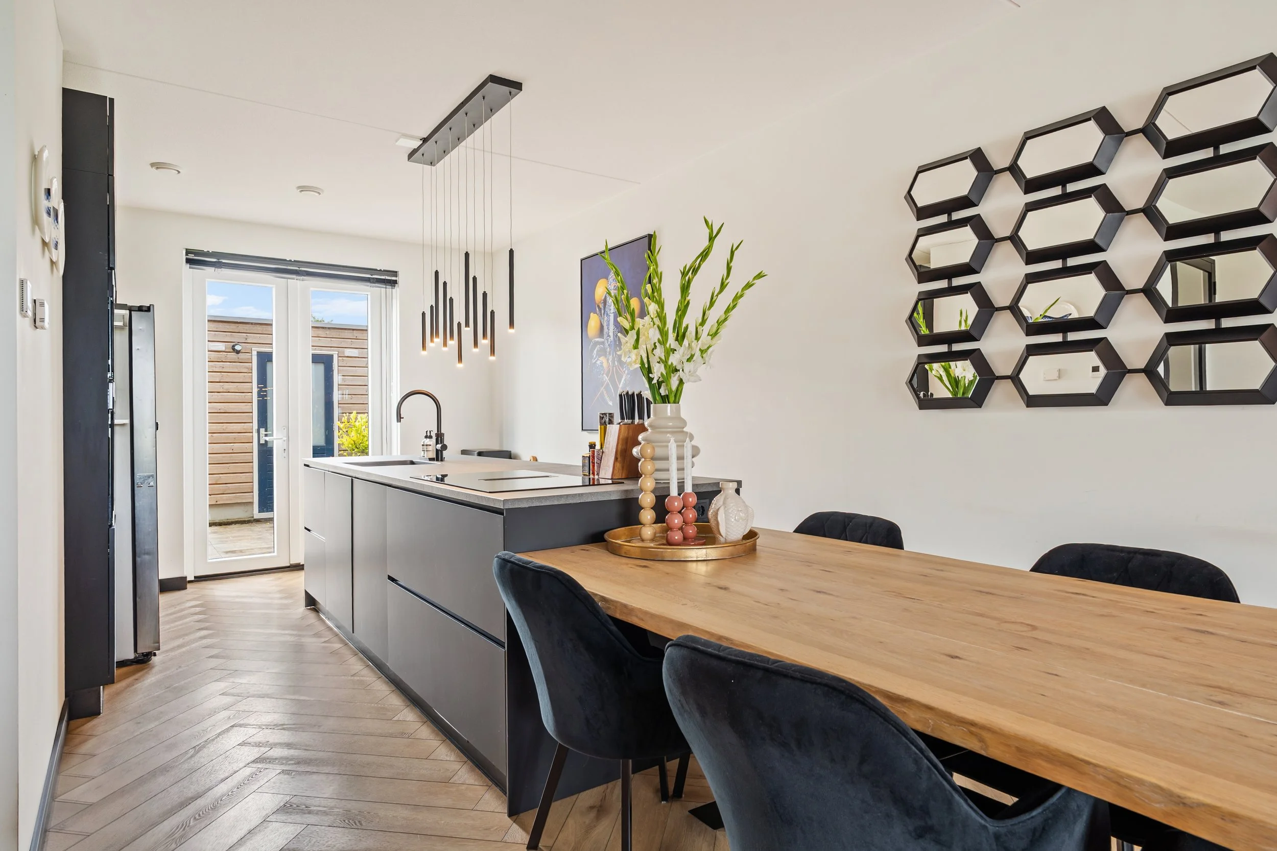 Een moderne keuken en eetkamer met een eettafel en vier stoelen, een hangende lamp, een vazenarrangement met witte bloemen, muurdecoraties met spiegelsegmenten en een deur naar een buitenterras.