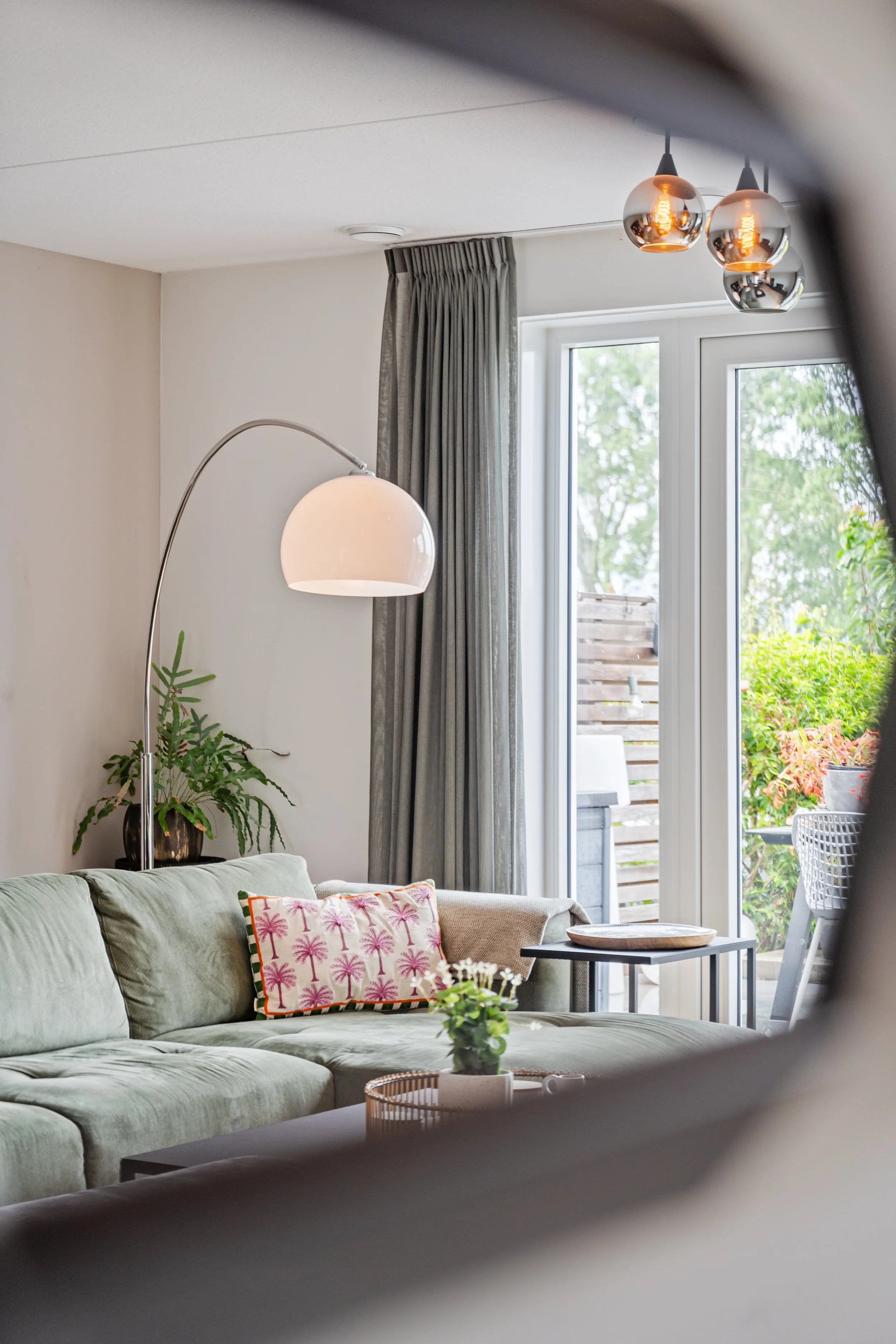 Een moderne woonkamer met een groene bank, roze-palm print kussen, binnenplant, metalen salontafel, grote raampartij en badkamer buiten te zien.