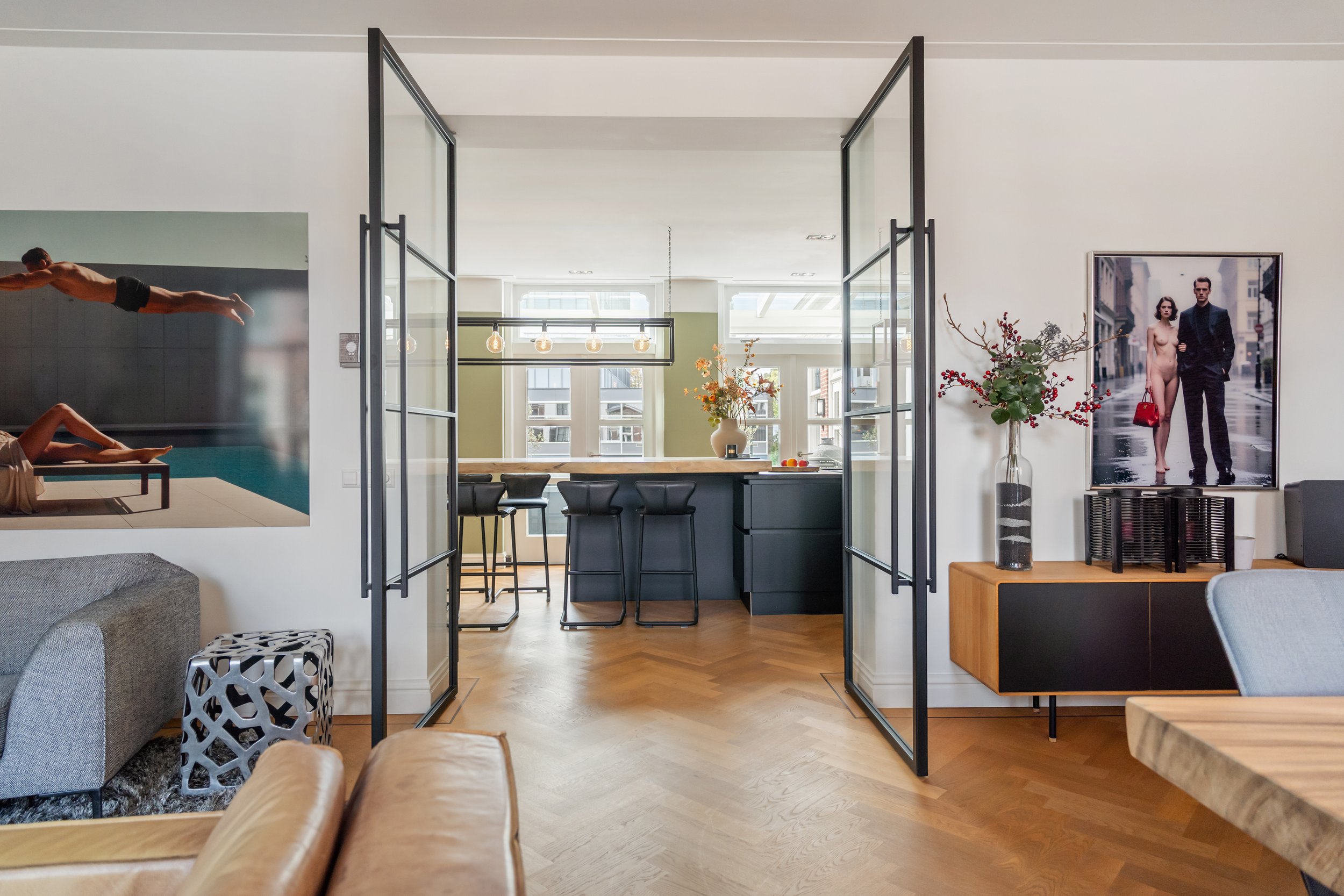 Interieur van een modern appartement met een open keuken en zithoek, grote ramen, bloemenvaas, televisie met een foto van een paar in formele kleding, en decoratieve elementen.