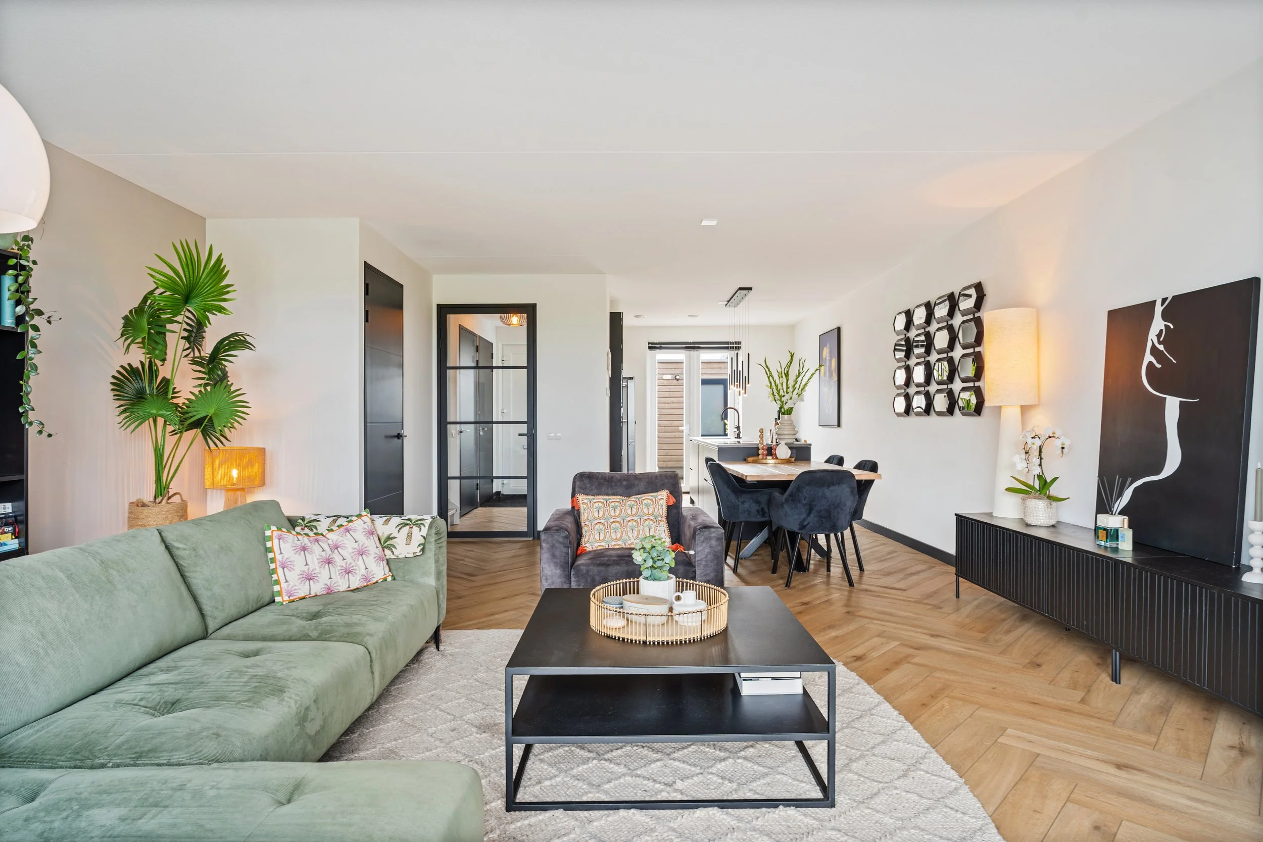Interieur van een woonkamer met een groene bank, een zwarte salontafel, een bank met patronen, planten, wanddecoraties en een eettafel met stoelen in een moderne, lichte woning.