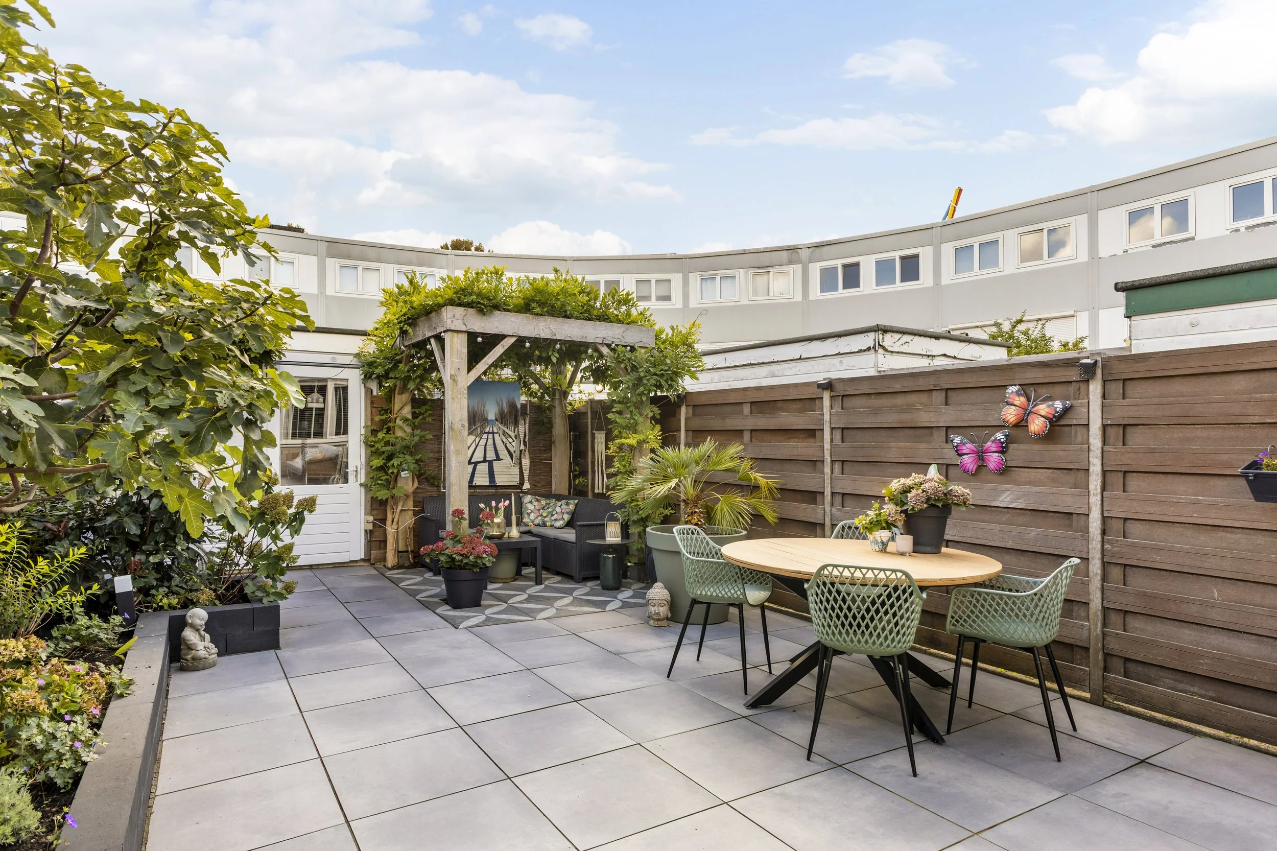 Een kleurrijke tuin met patio, meubels, bloemen, planten, tuinbeeld, vlinders, en een houten hek, onder een blauwe lucht met wolken.