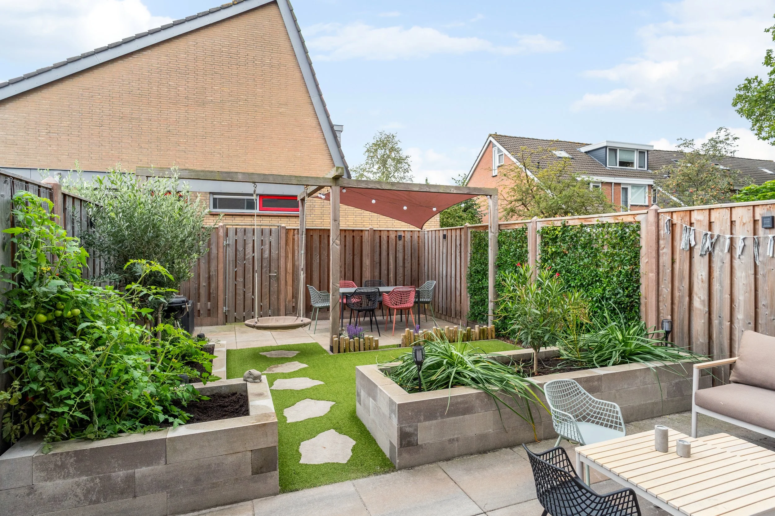 Een tuin met een houten omheining, zitplaatsen, een tuinbed met groene planten en een grasstrook met stenen, omgeven door huizen en bomen onder een licht bewolkte hemel.