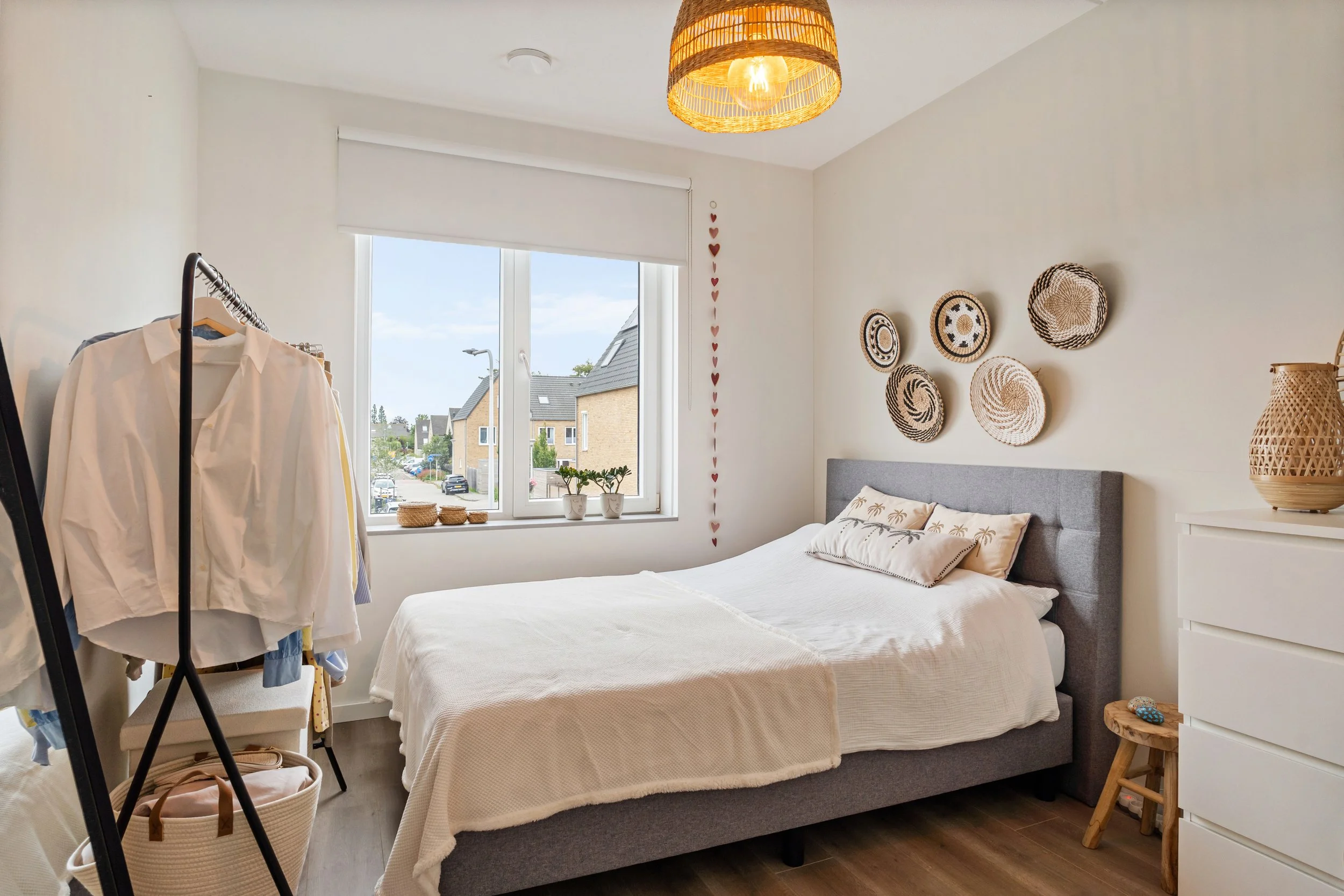 Een slaapkamer met een grote bed, raam met uitzicht op huizen, wanddecoratie met manden en een plafondlamp van rotan.