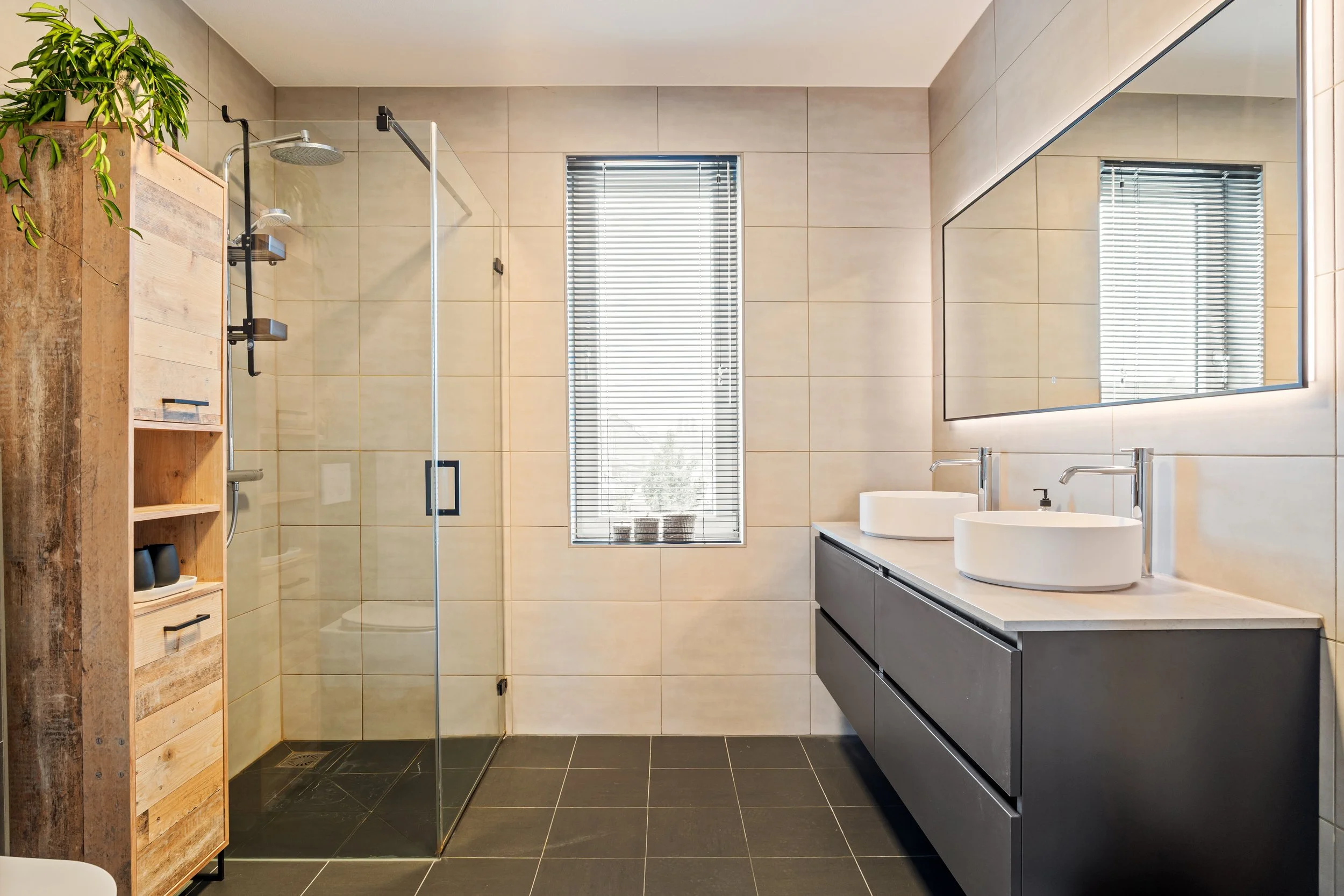 Moderne badkamer met inloopdouche met glazen deur, twee wastafels met badkamerspiegels, grote ramen met jaloezieën, en een houten kast met planten.